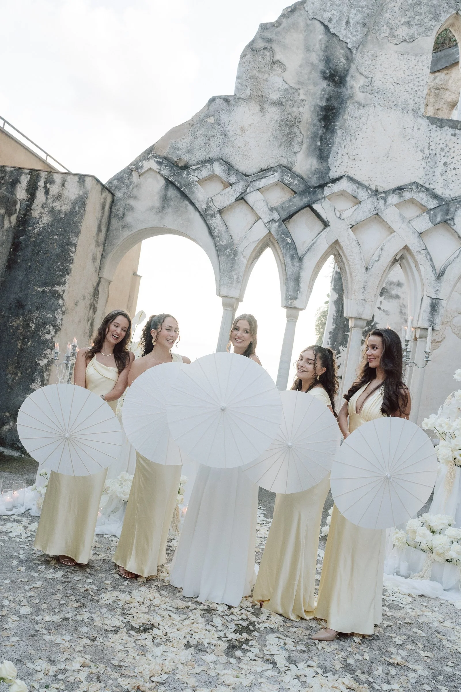 Women in cream-colored dresses holding white parasols and smiling, at a wedding or celebration outdoors with an old stone architectural structure in the background.