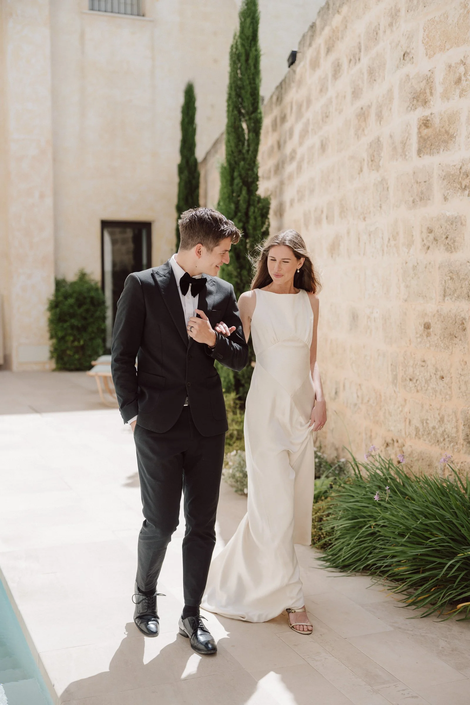 A man in a black tuxedo and a woman in a white wedding dress walking together outdoors near a stone wall with tall cypress trees in the background.