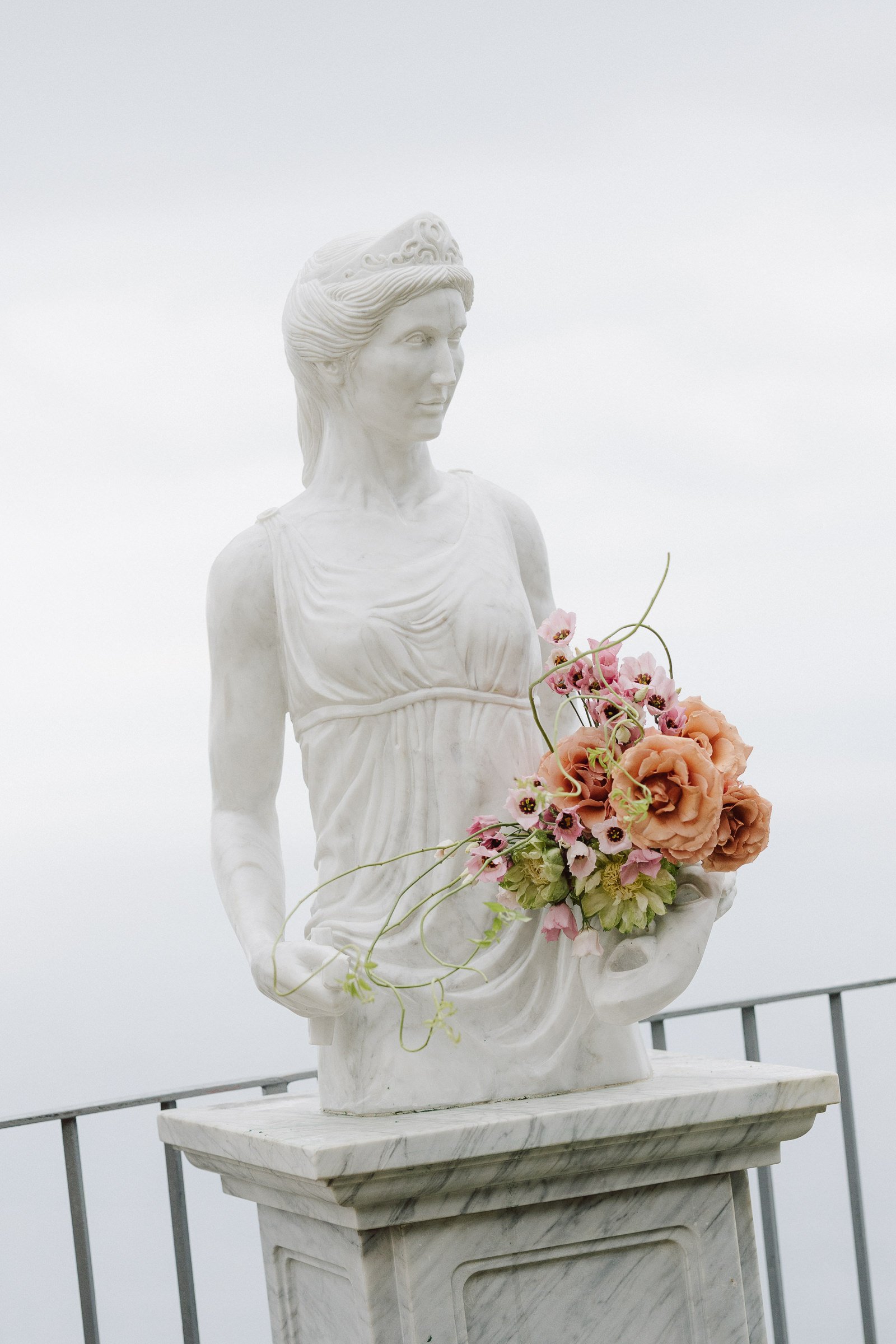 A white marble statue of a woman with flowers in her woven arm, standing on a marble pedestal against a foggy sky background.