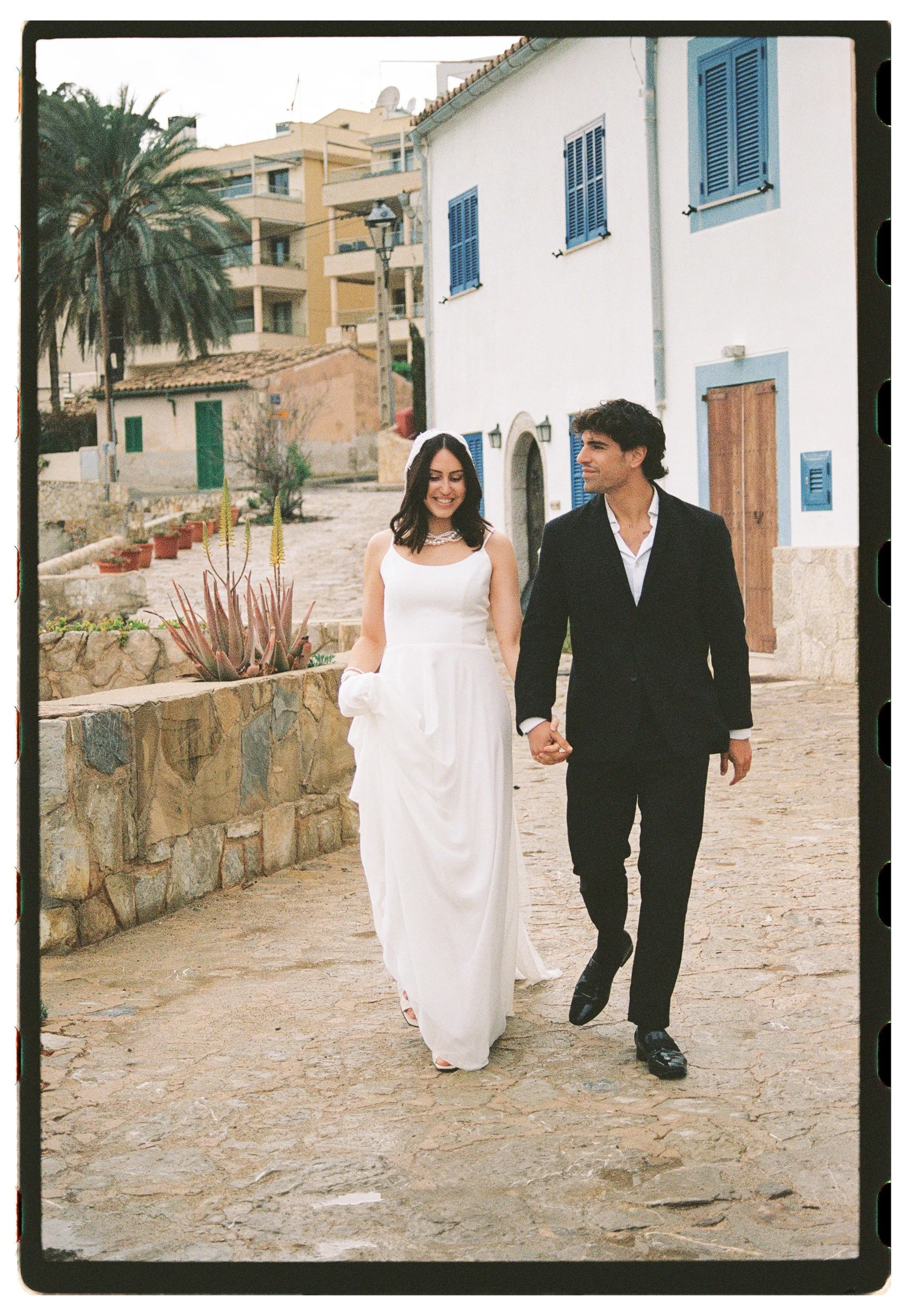 A couple dressed in wedding attire holding hands and walking along a cobblestone street in a Mediterranean town with white and blue buildings, potted plants, and a palm tree.
