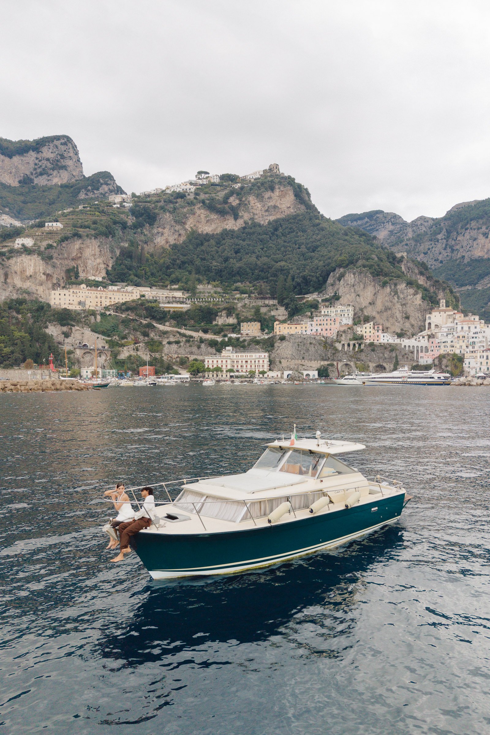 A yacht with two people sitting at the bow, moving through a harbor with coastal mountains and buildings in the background.