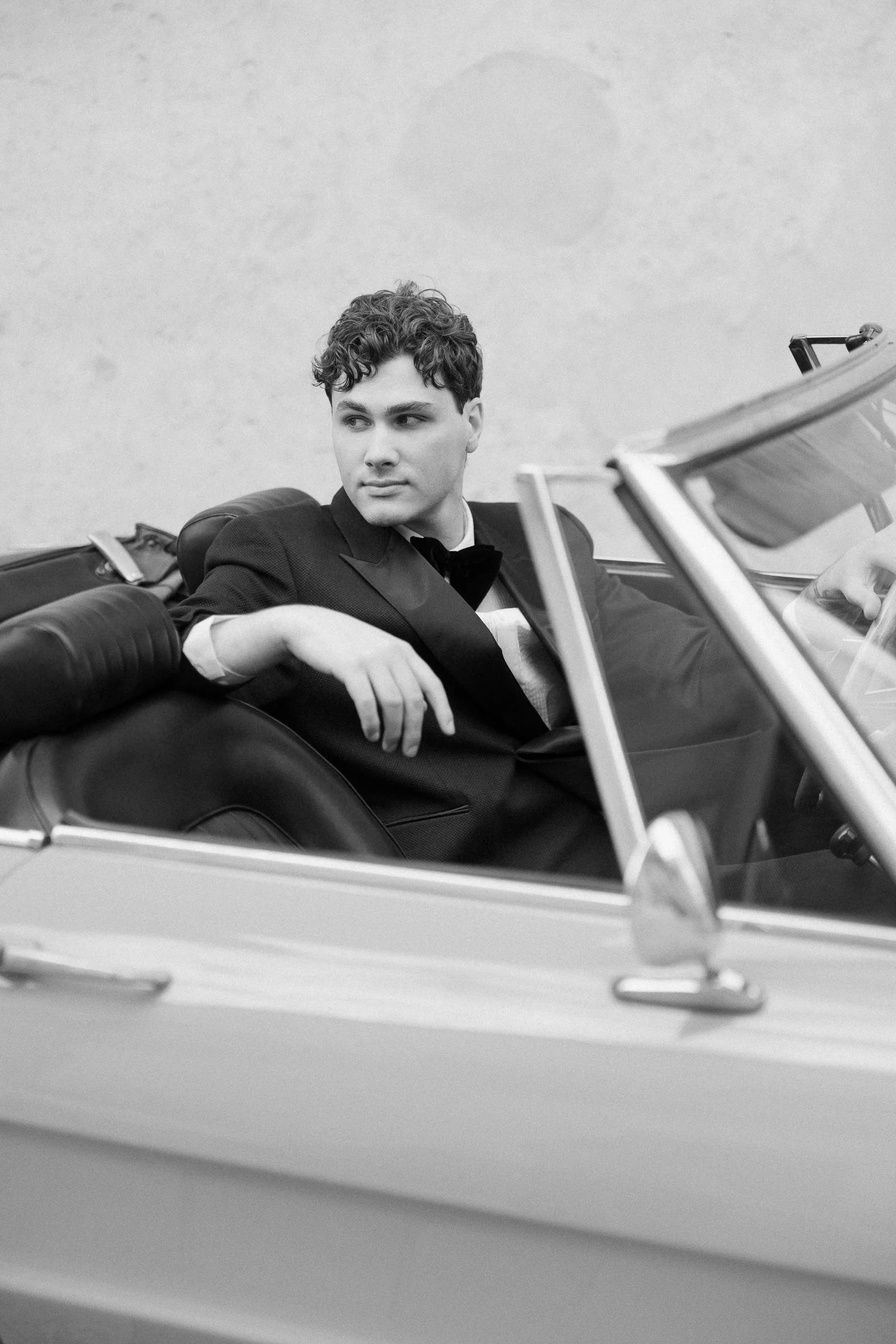 Black and white photo of a young man in a tuxedo sitting in a vintage convertible car, looking to his right with a relaxed pose.