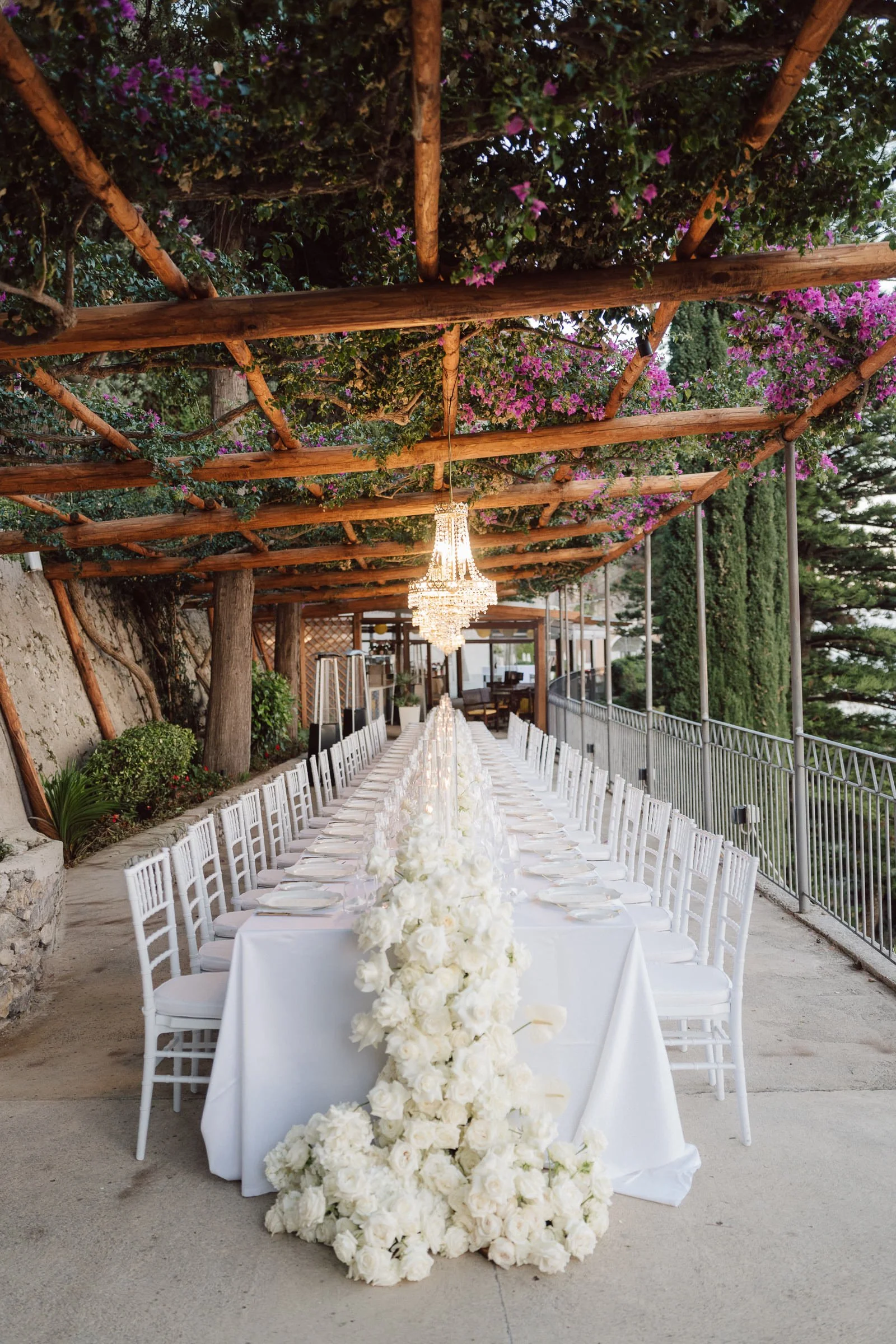 Elegant outdoor banquet setting with a long white table surrounded by white chairs, decorated with a tall floral centerpiece of white roses and flowers. Above, a wooden pergola draped with purple flowering vines and a chandelier hanging in the center