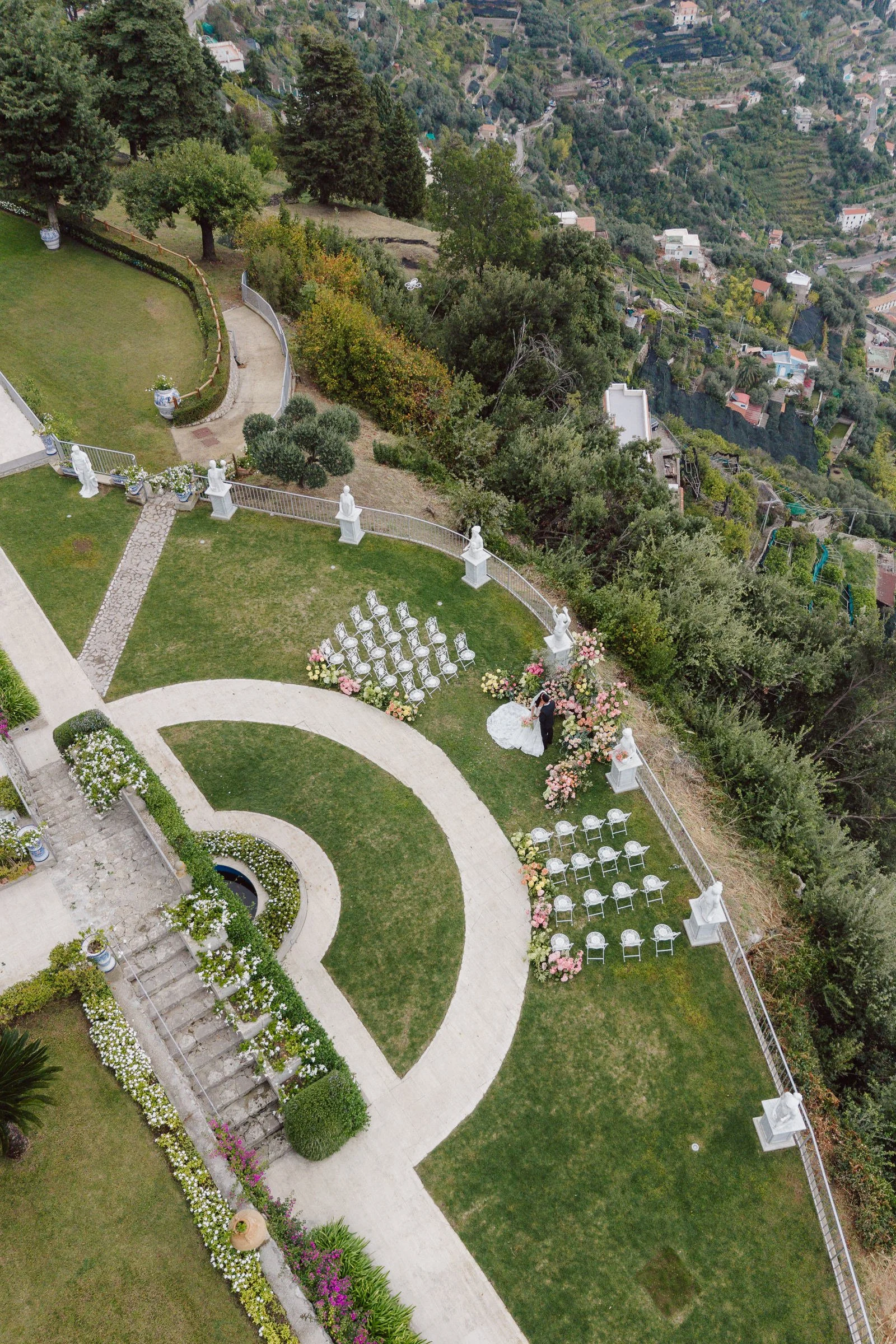 A wedding ceremony setup on a hilltop overlooking a green hillside with houses. There are white chairs arranged for guests, statues, flowers, and a bride and groom standing under a floral arch.