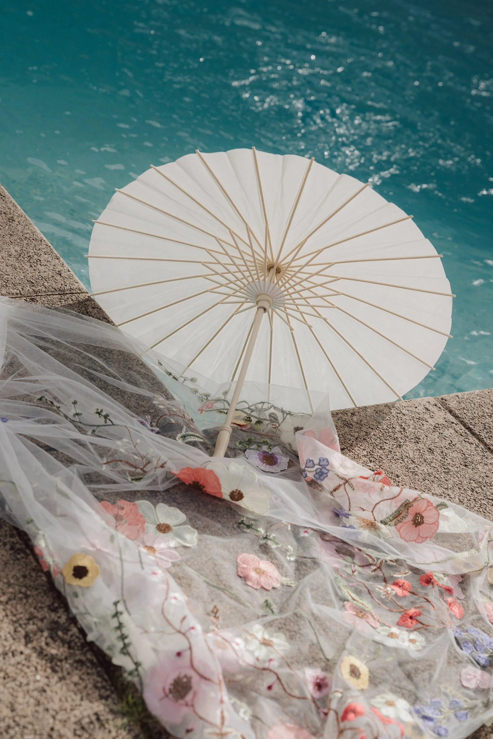 A white paper parasol icon is placed on a stone ledge next to a swimming pool, surrounded by floral embroidered fabric.