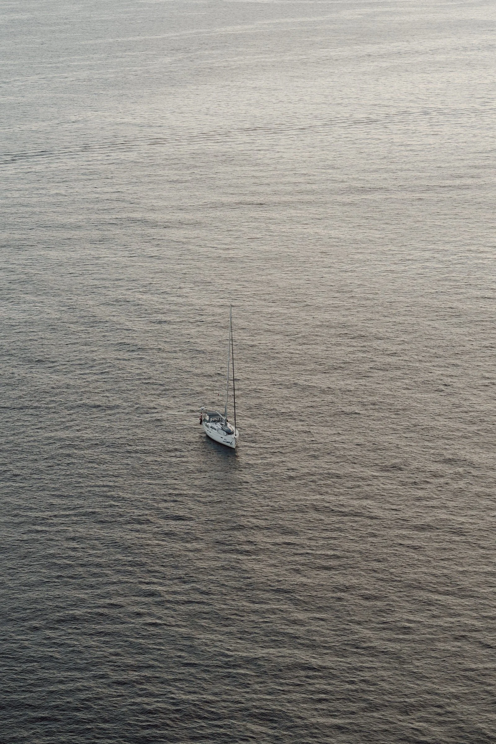 A small white sailboat floating alone on a calm, expansive body of water, possibly a lake or ocean, at dusk or dawn.