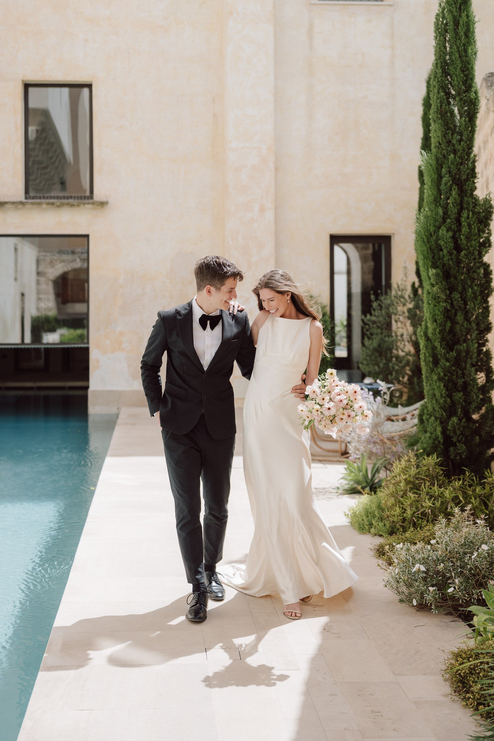 A smiling bride and groom walking arm in arm near a swimming pool during a wedding celebration, with a modern beige building and tall green trees in the background.