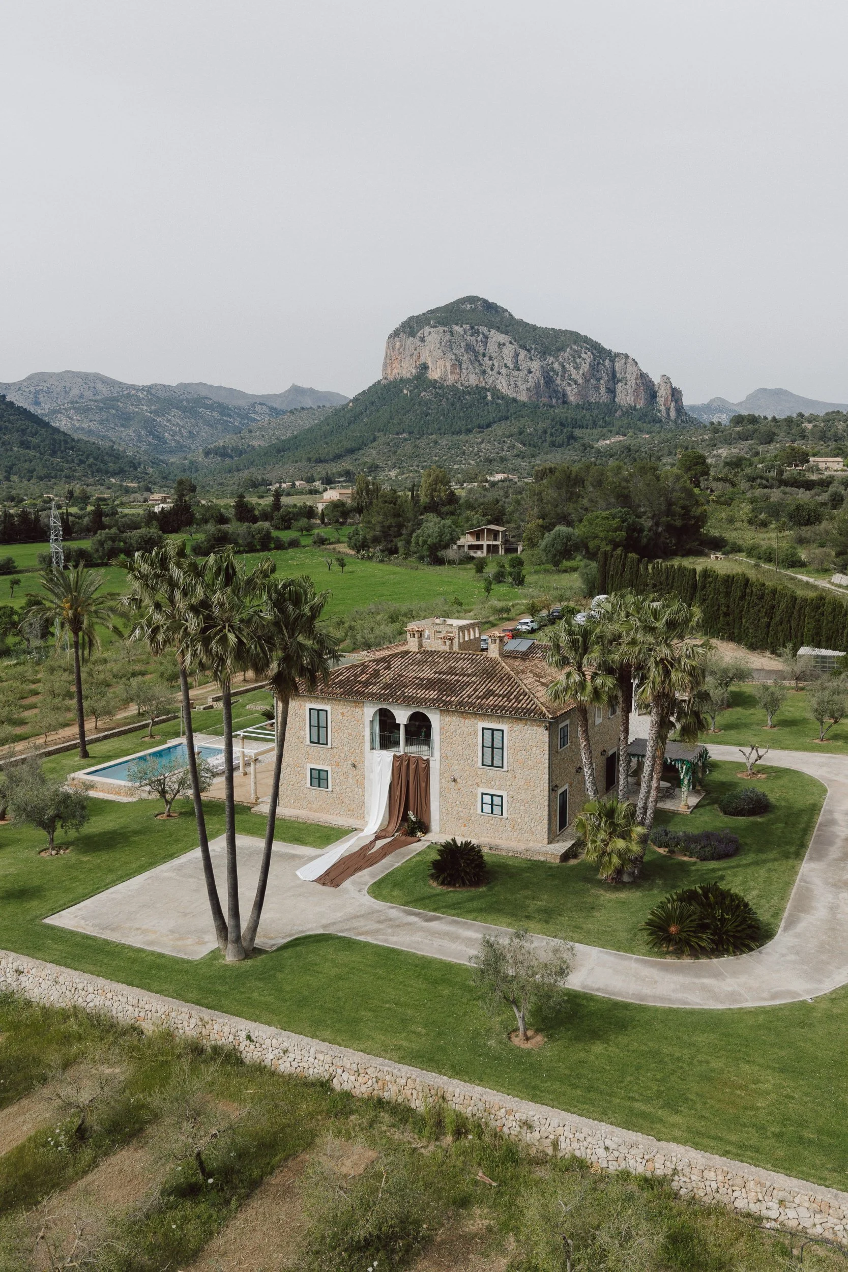 A house with a stone exterior and a tiled roof, featuring a brown and white draped outdoor canopy extending from a balcony, is surrounded by a well-maintained garden with tall palm trees, smaller trees, bushes, and a path leading up to the house. The