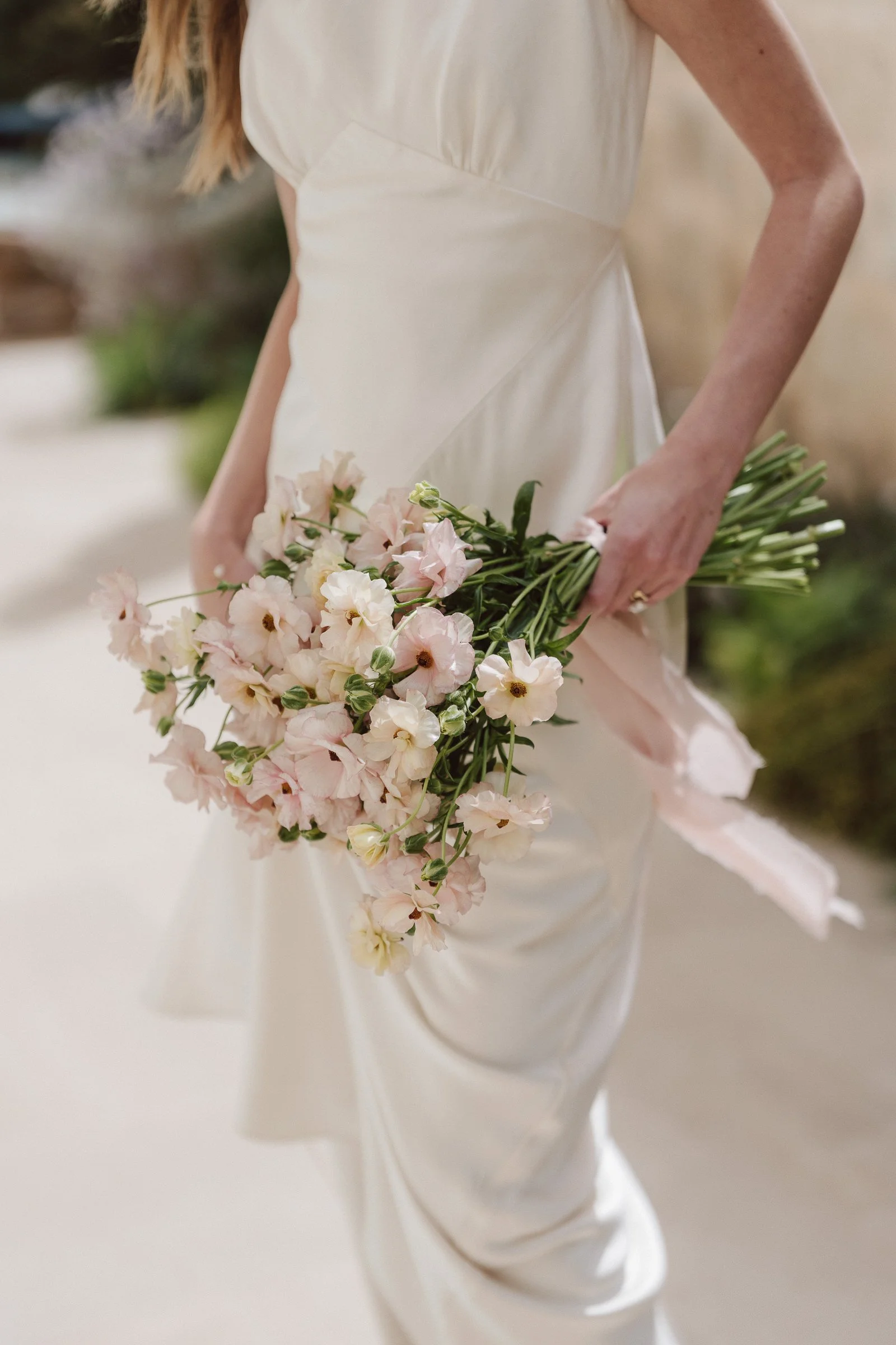A person in a cream-colored dress holding a bouquet of light pink flowers with green stems, standing in an outdoor setting.