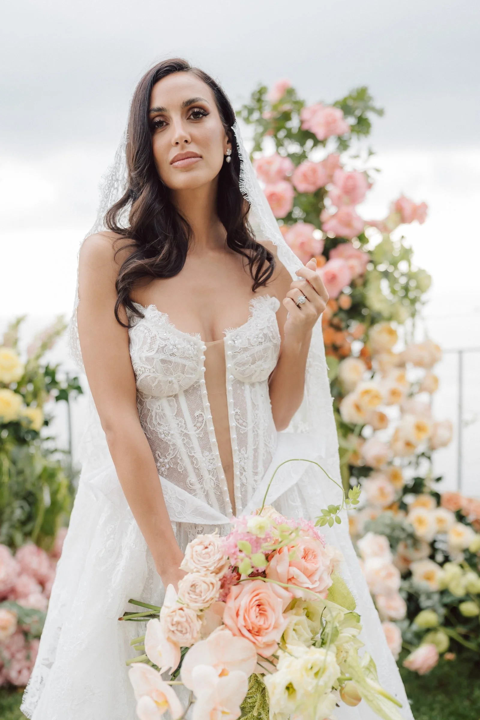 A woman in a white wedding dress holding a bouquet of pink and white flowers outdoors with pink and peach flowers in the background.