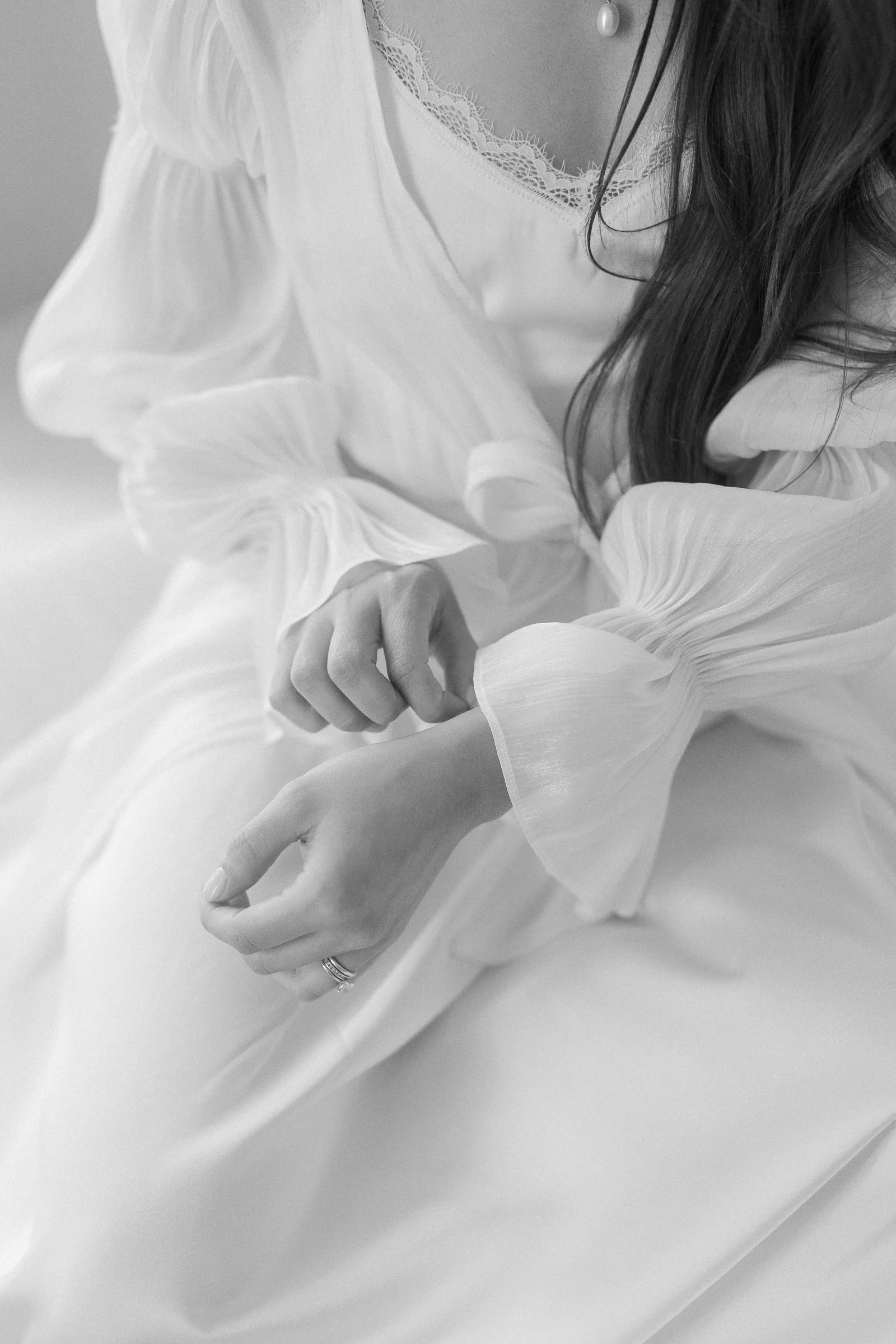 Black and white photograph of a person with long dark hair wearing a light-colored dress with lace detail on the neckline. The person's hands are touching, with one hand holding the other, and they are wearing a ring.