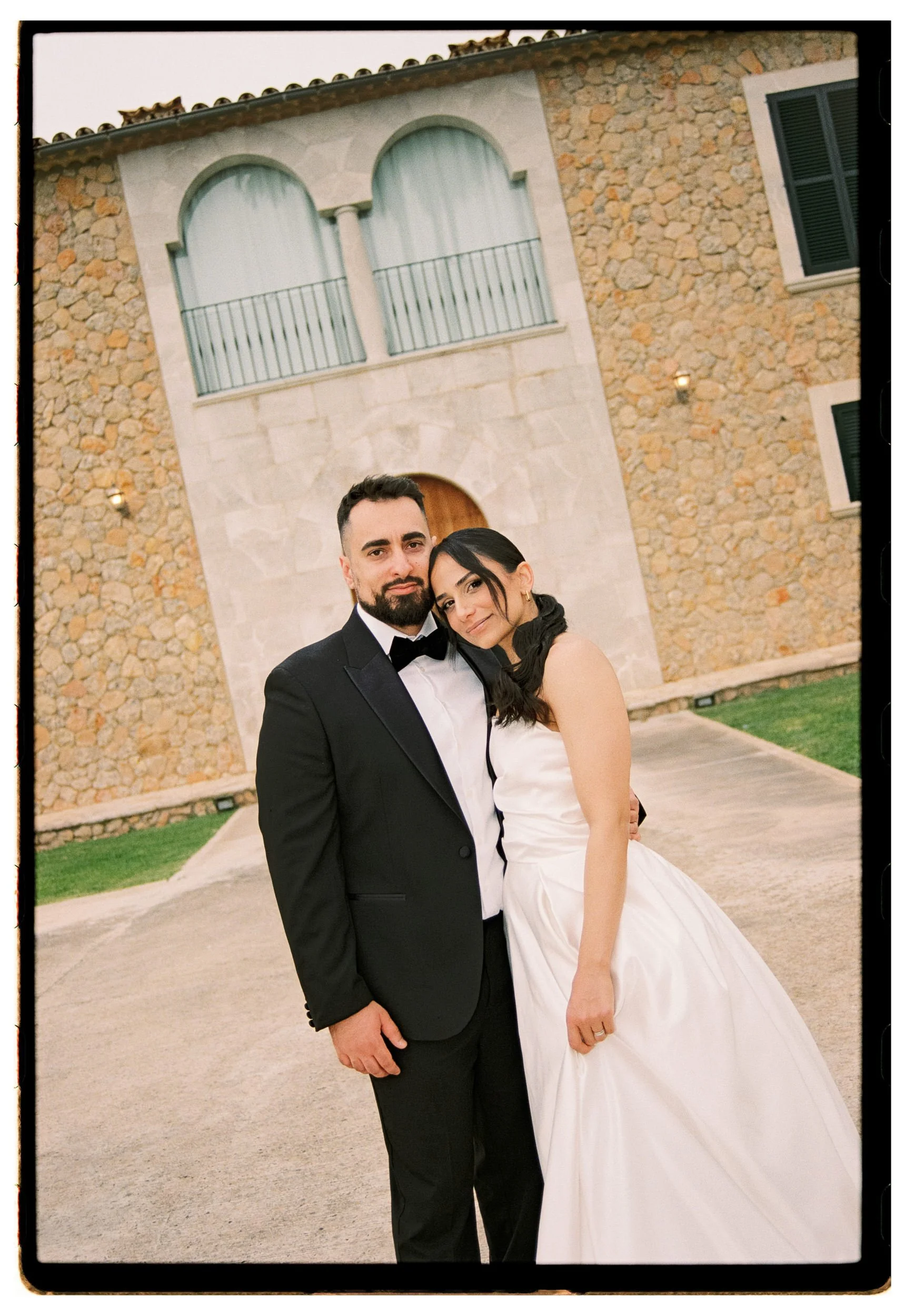 A newlywed couple in wedding attire stands close together outdoors in front of a stone building with an arched window and black shutters.