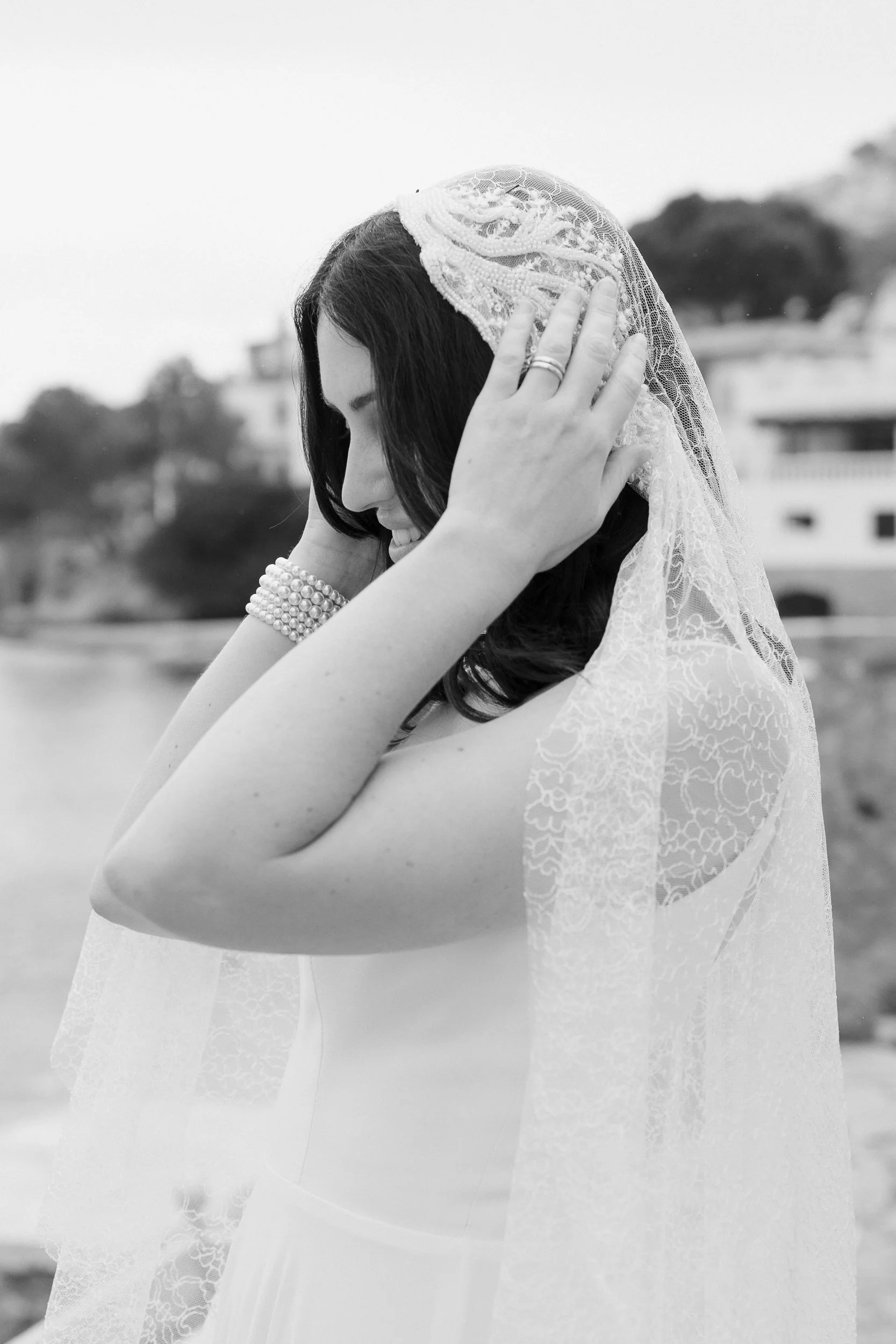 A woman in a wedding dress and lace veil smiling, with her hands near her head and wearing pearl bracelets and rings, outdoors with blurred trees and buildings in the background.