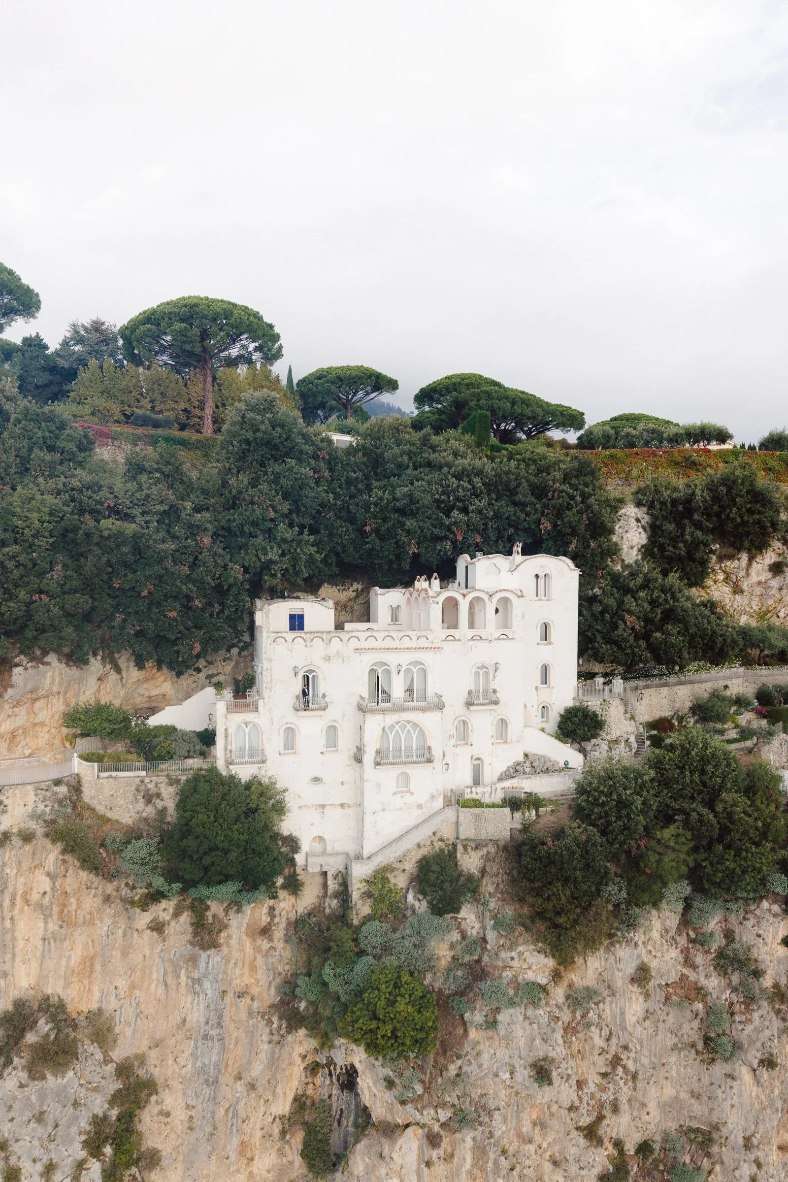 A scenic view of a hillside with white Mediterranean-style buildings surrounded by lush green trees and shrubs, with a cloudy sky overhead.