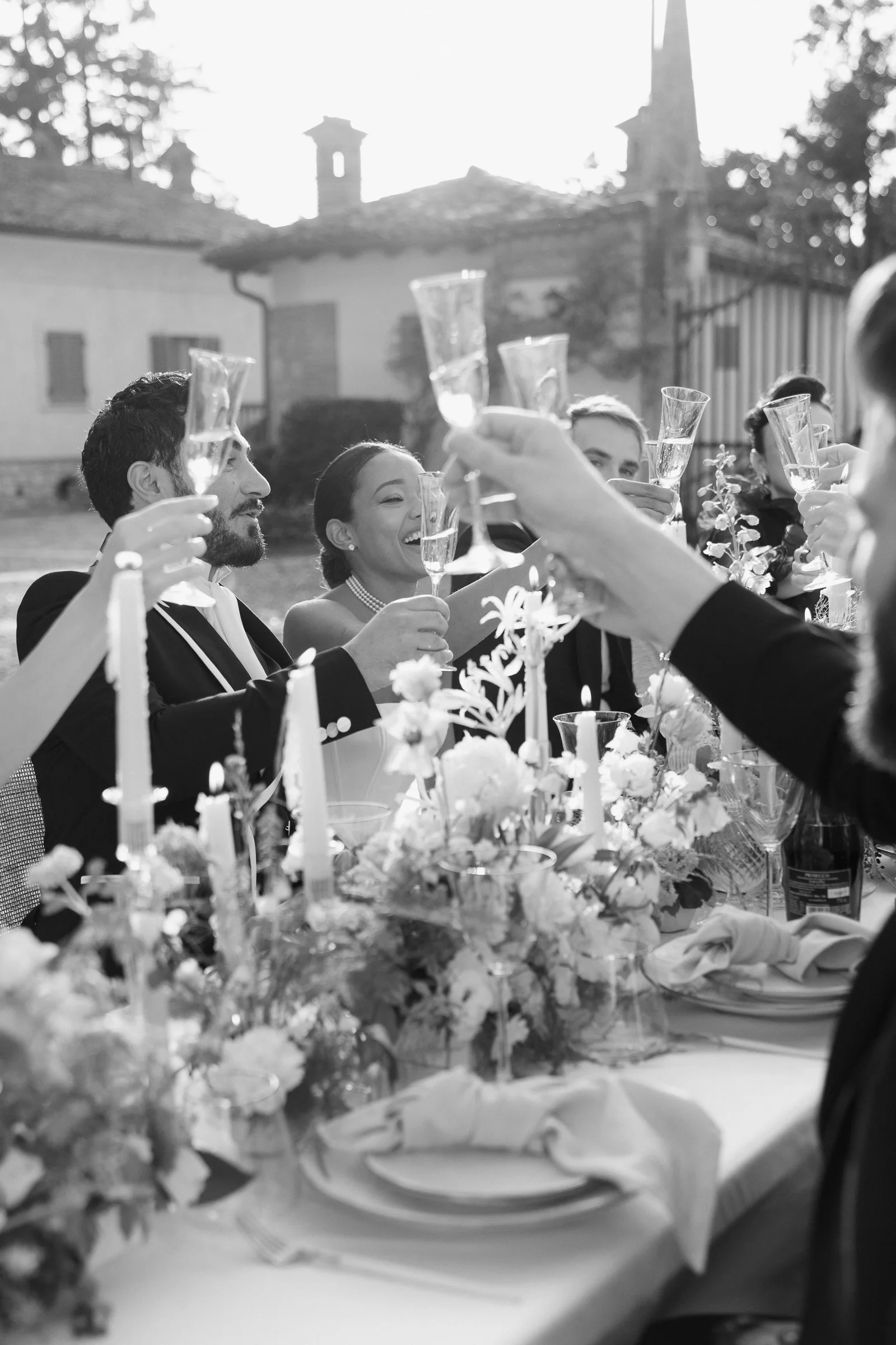 People celebrating with champagne glasses at an outdoor wedding reception, with a decorated table and floral arrangements visible.