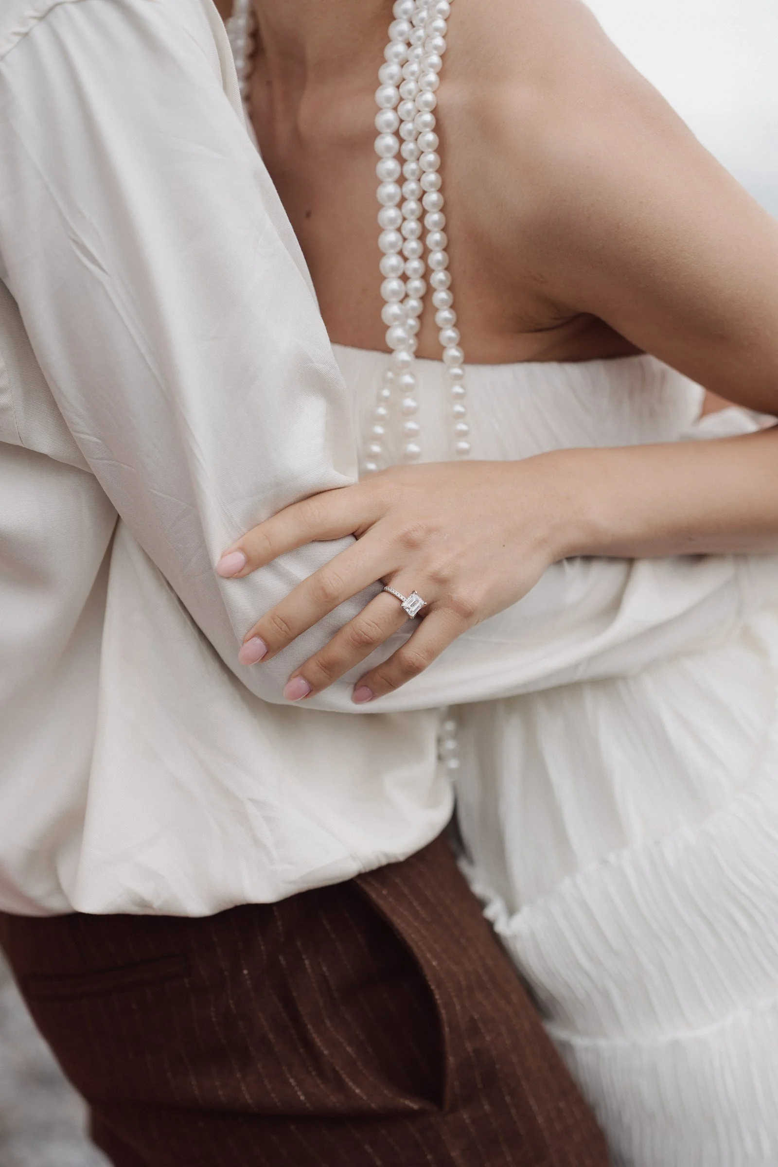 Close-up of a woman showing her shoulder, wearing a pearl necklace, a white satin top, and a diamond ring on her finger. She is also wearing brown and white striped pants.