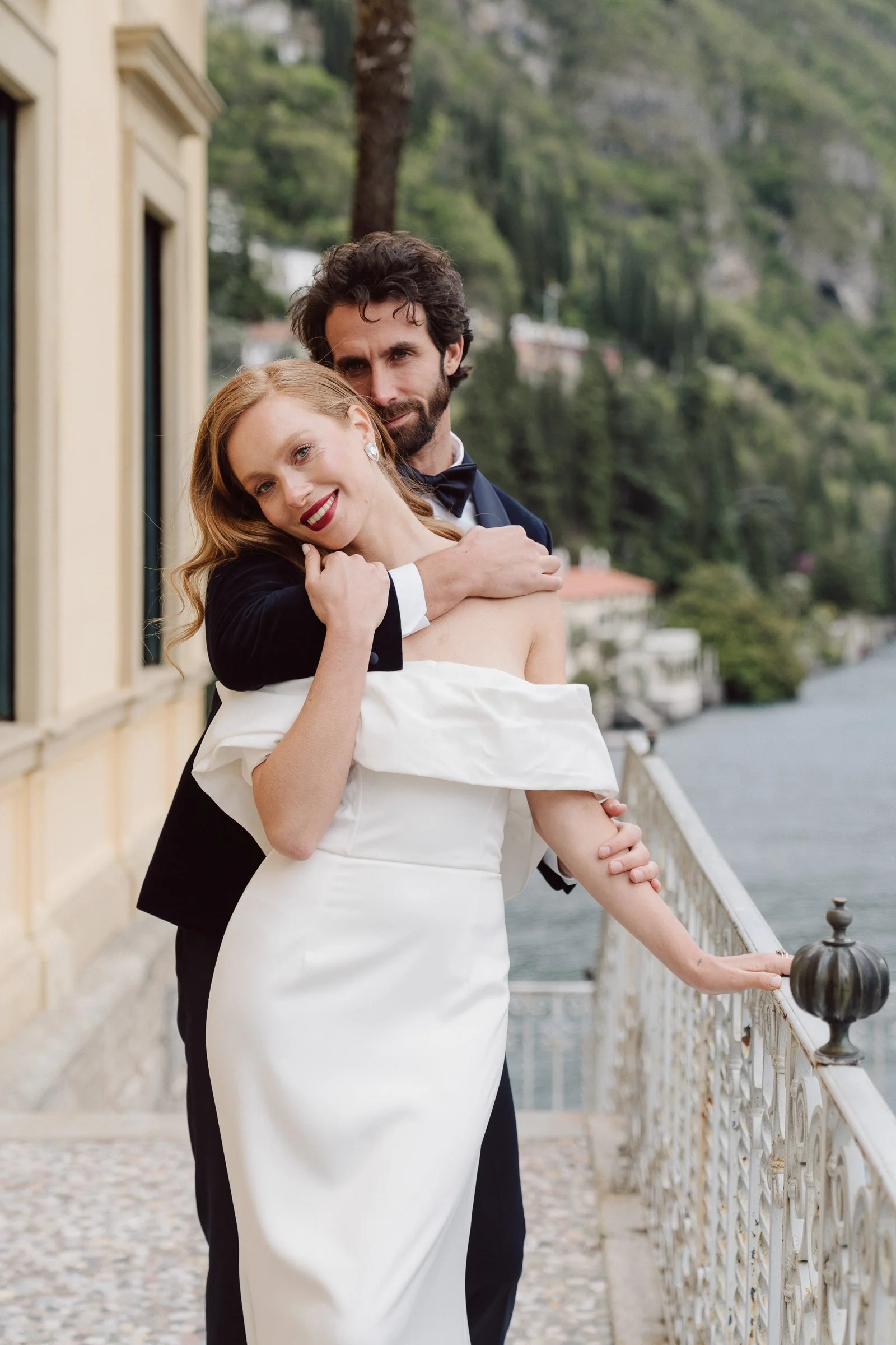 A smiling woman in a white strapless dress and earrings is embracing a man in a black tuxedo with a bowtie on a waterfront promenade, with greenery and buildings in the background.