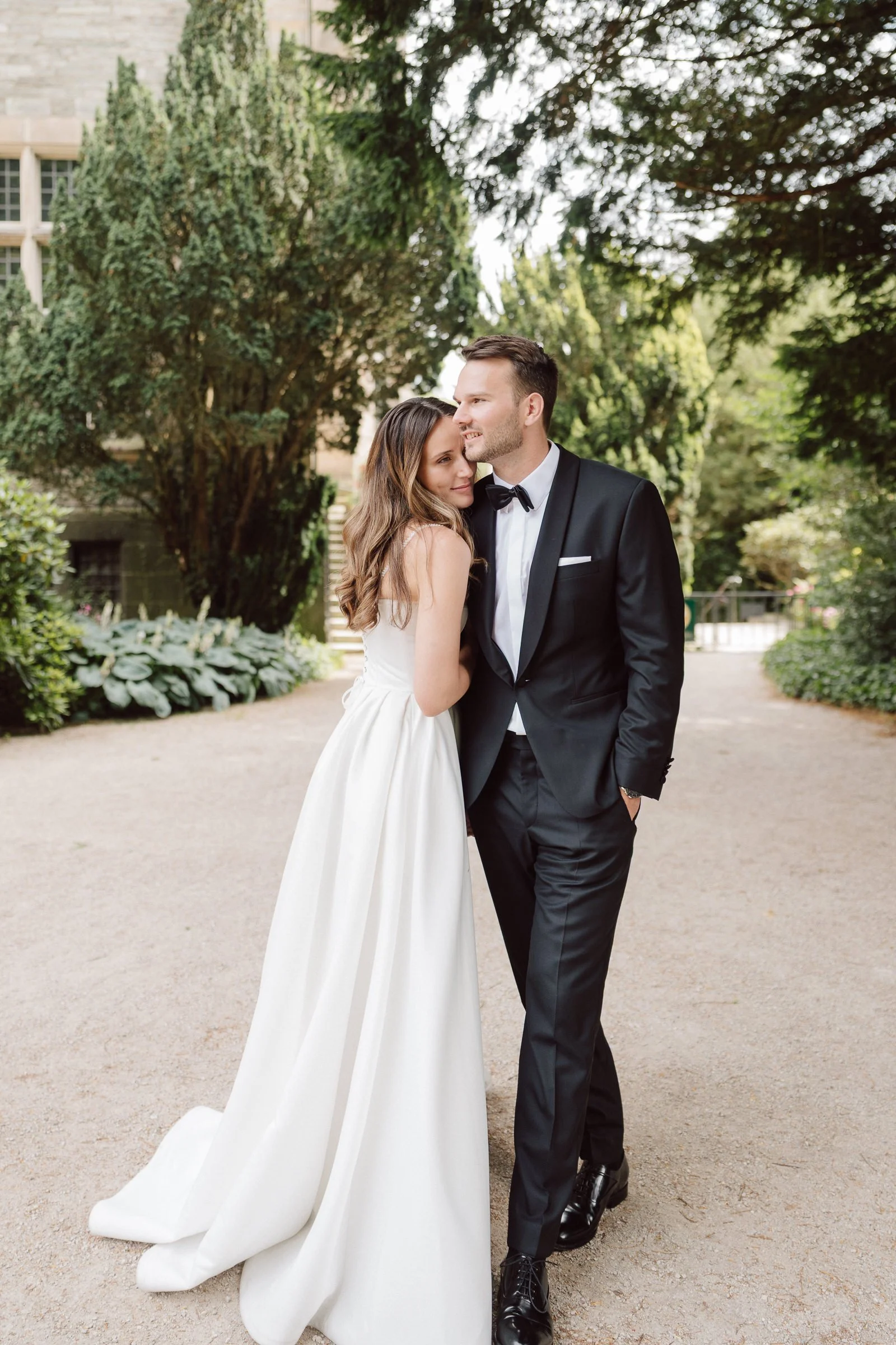 A bride and groom standing close together outdoors in a garden, with the bride wearing a white wedding dress and the groom in a black tuxedo, embracing each other and smiling.