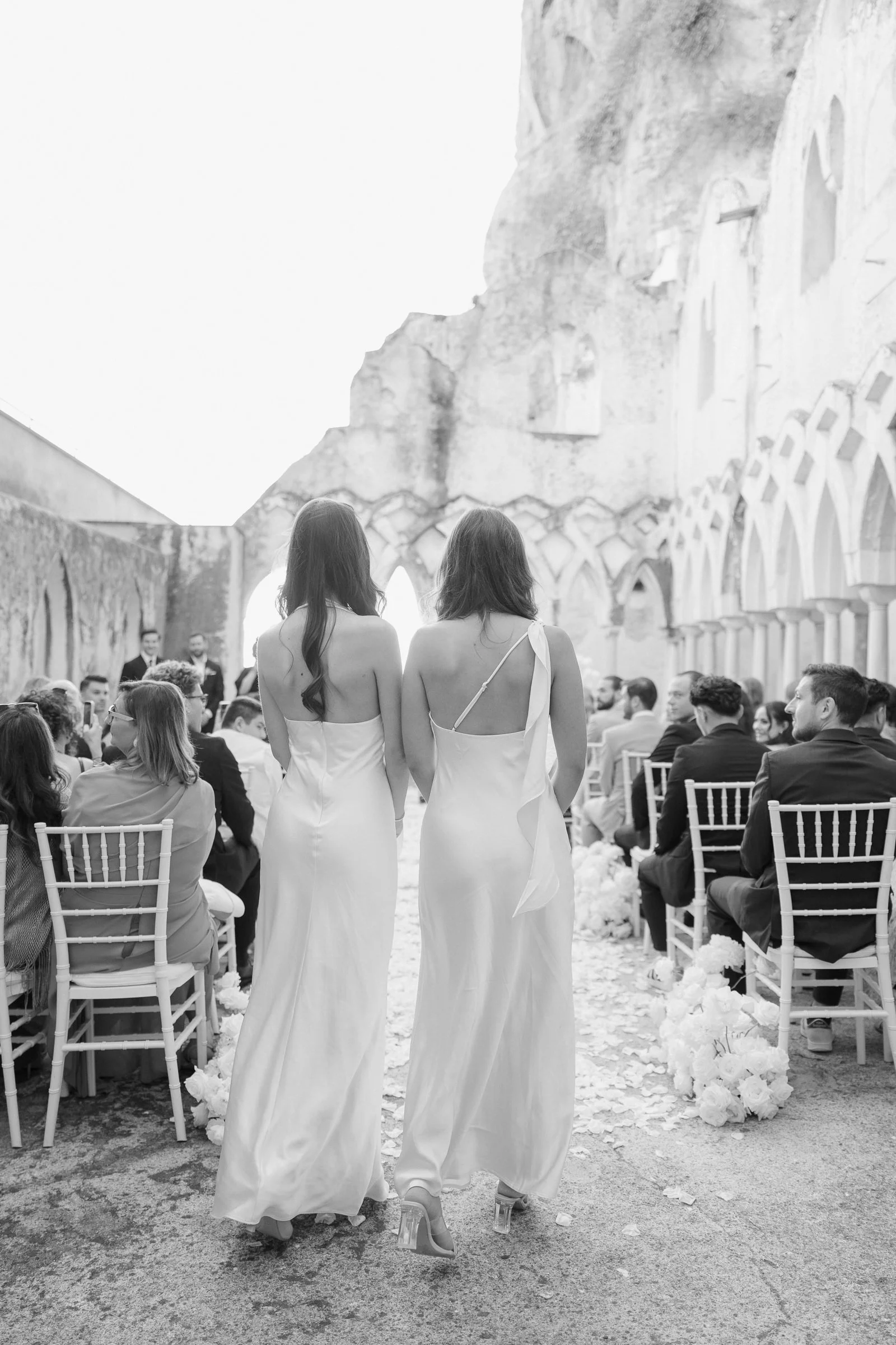 Two brides in white wedding dresses walking down the aisle at a wedding ceremony. Guests seated on either side of the aisle, with a stone wall and decorative arches in the background. The photo is black and white.