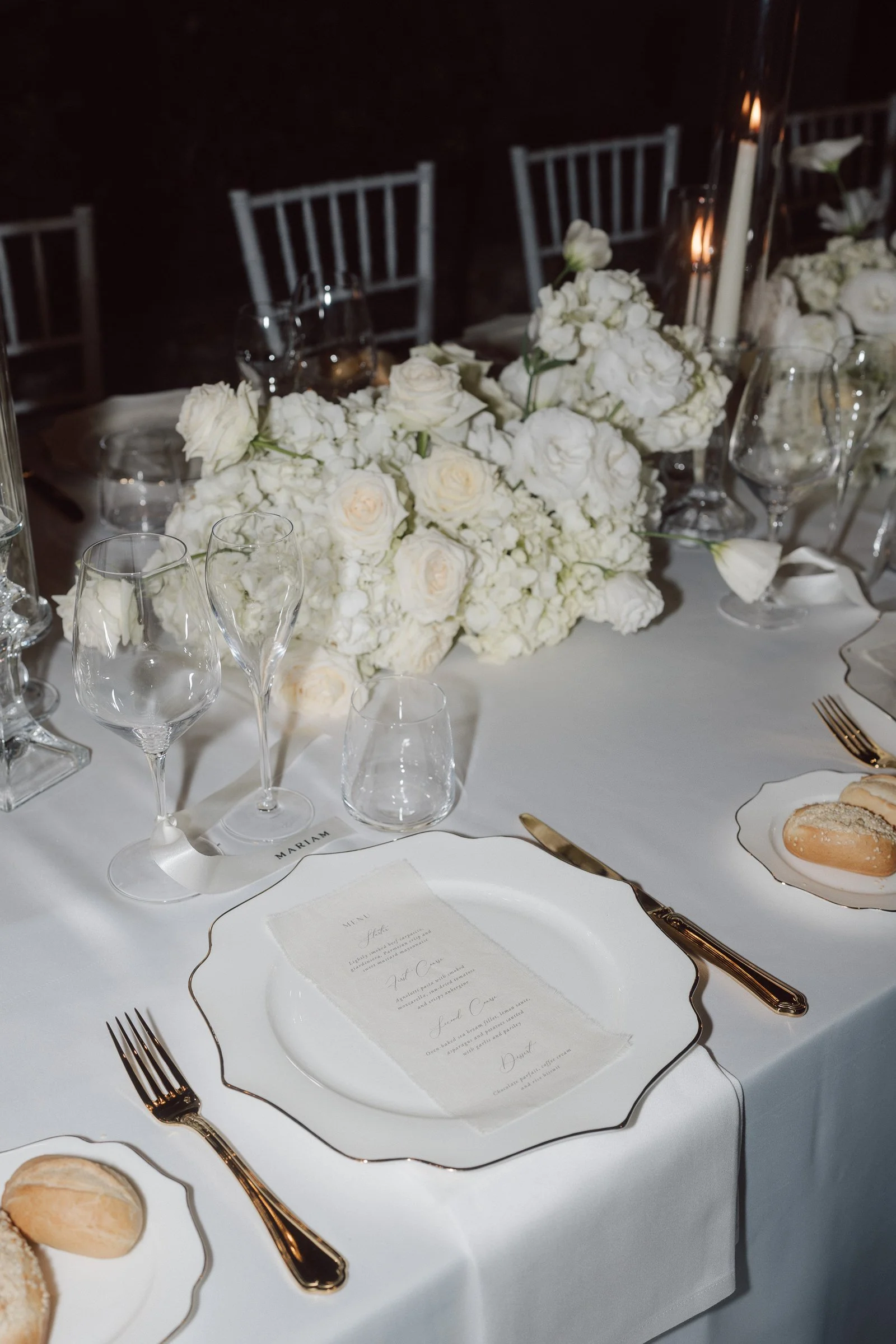 Elegant dinner table setting with white floral centerpiece, wine glasses, candles, and gold-cutlery