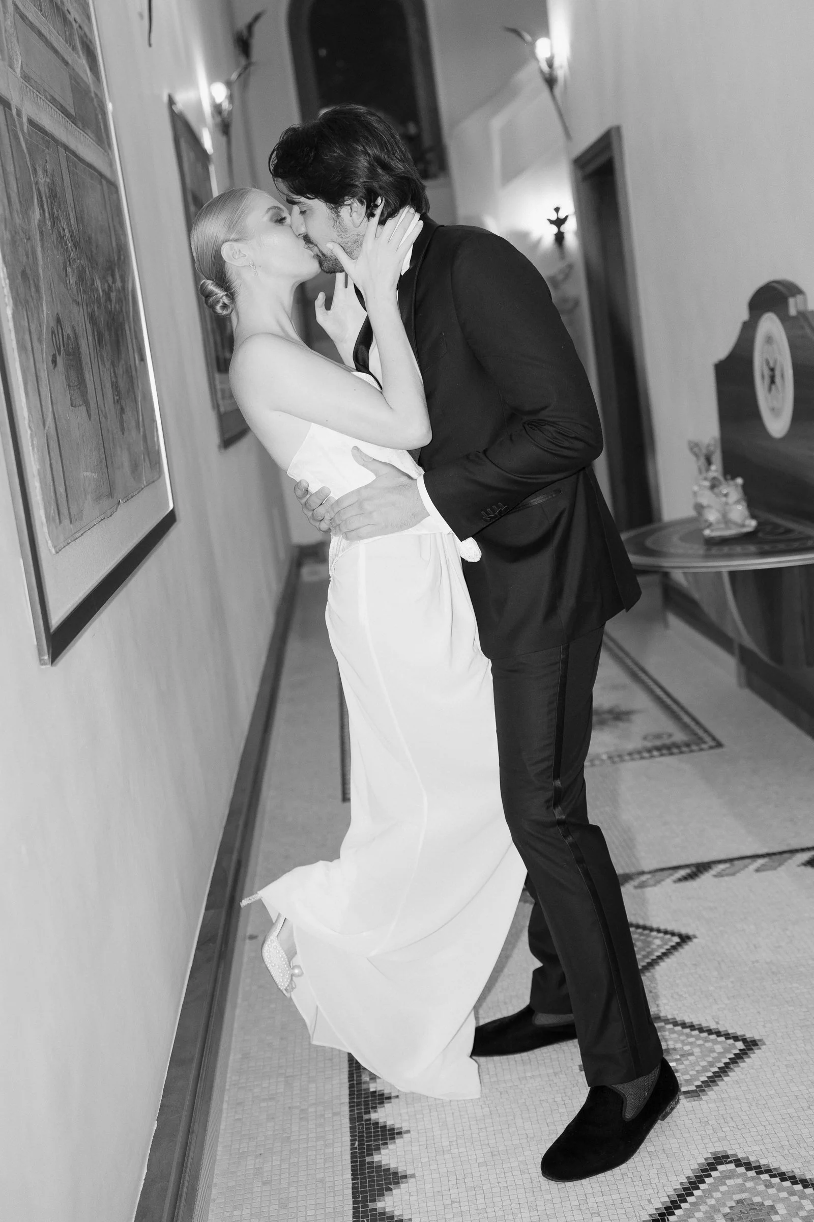 A man and woman kissing, dressed in wedding attire, in an indoor hallway with framed pictures on the wall and decorative light fixtures.