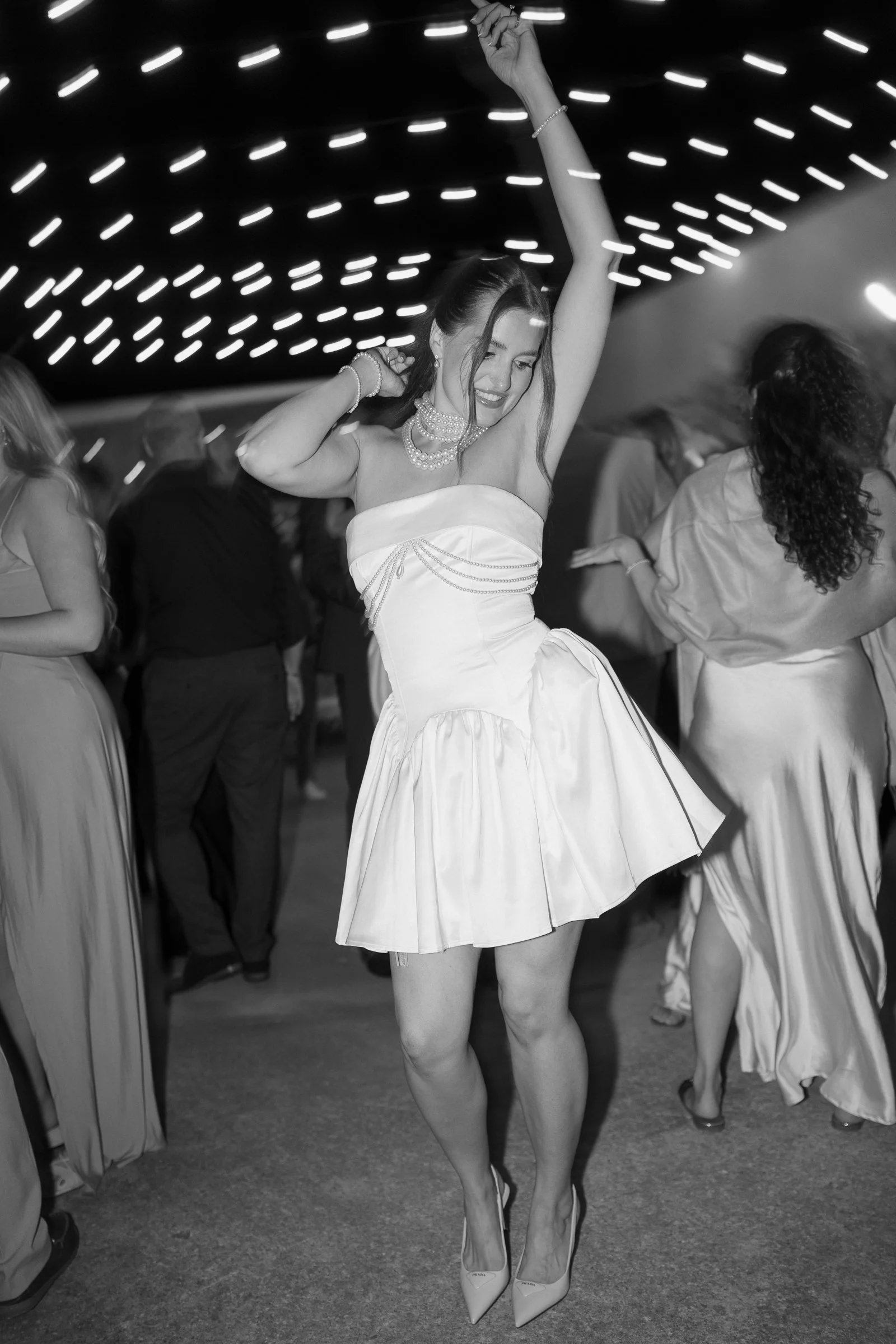 A woman in a strapless dress and high heels dancing at a party or dance event, surrounded by other people.