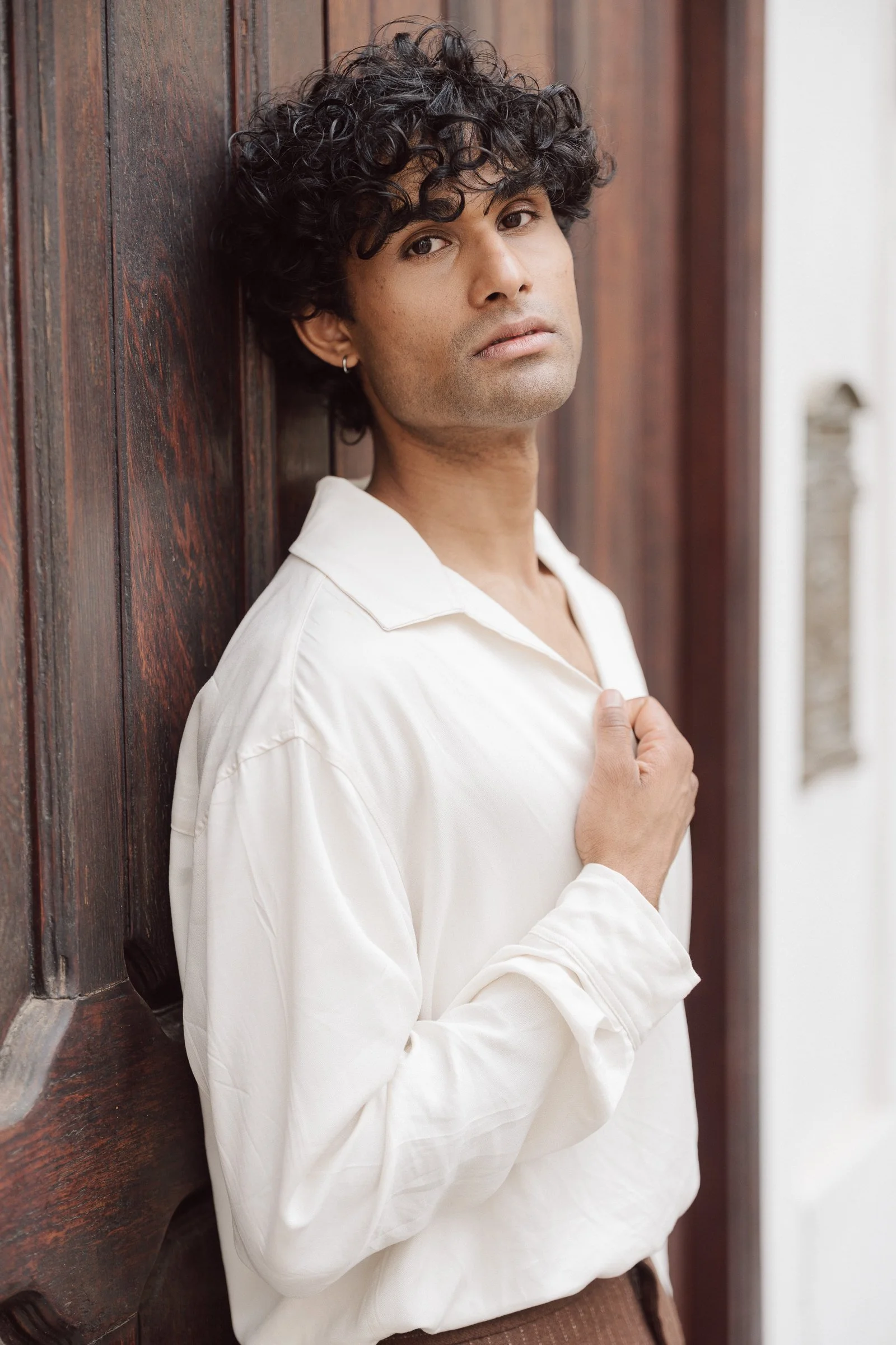A young man with curly dark hair, wearing a white shirt, leaning against a wooden wall, looking at the camera with a neutral expression.