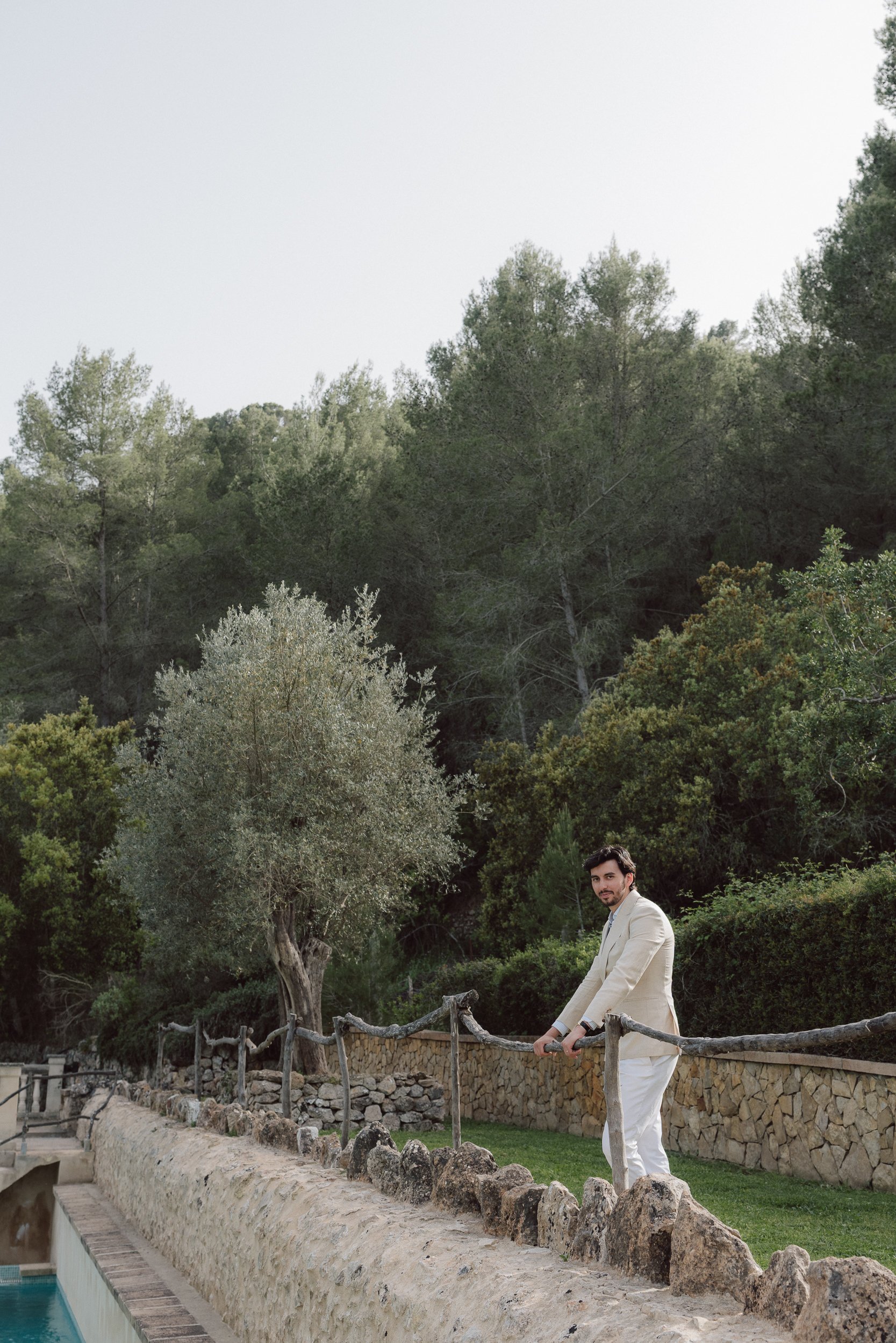A man in a white suit standing near a stone wall and wooden fence by a pool, with tall green trees and cloudy sky in the background.