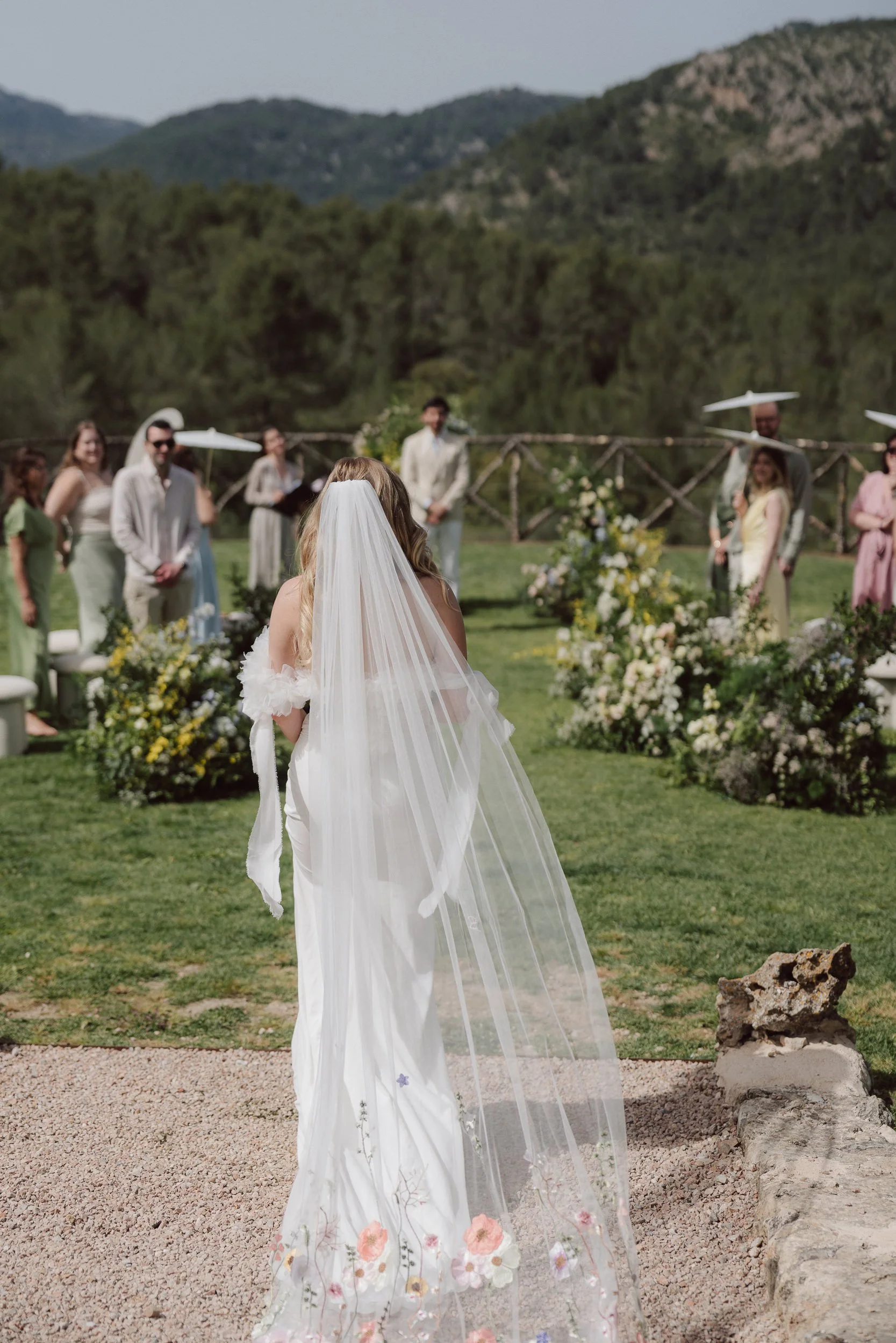 A bride with long blonde hair and a white veil stands outdoors during a wedding ceremony, facing away from the camera. She is dressed in a white gown with floral embroidery on the bottom. The scene is set in a scenic outdoor location with a grassy ar