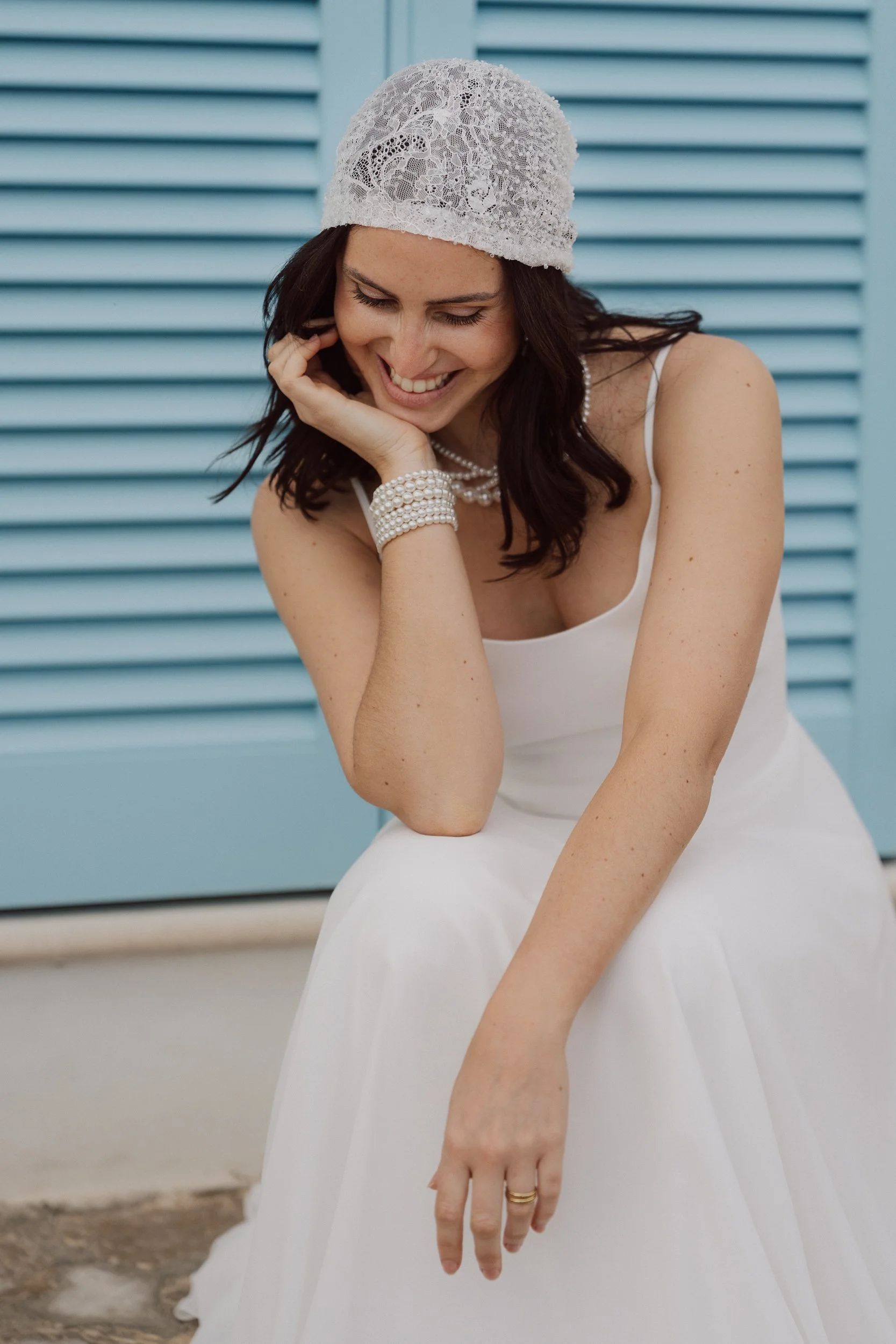 A woman in a white dress, lace head covering, and pearl jewelry is smiling while sitting outdoors in front of a blue louvered wall.