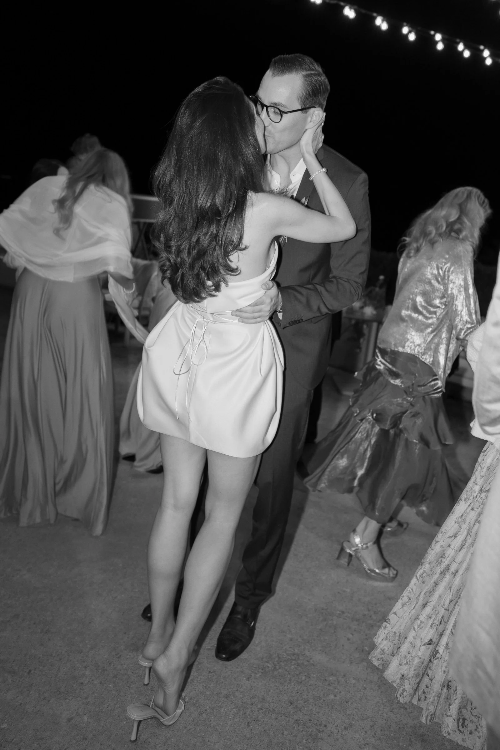 A couple sharing a kiss at a nighttime event, with string lights overhead and women in fancy dresses dancing in the background.