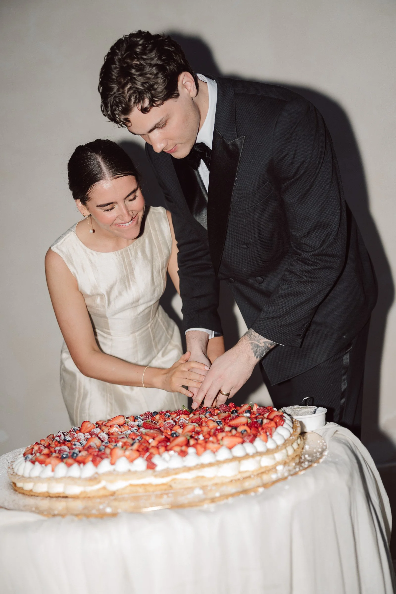 A man and a woman in formal attire cutting a large decorated cake with strawberries, blueberries, and whipped cream layers, at a celebration.