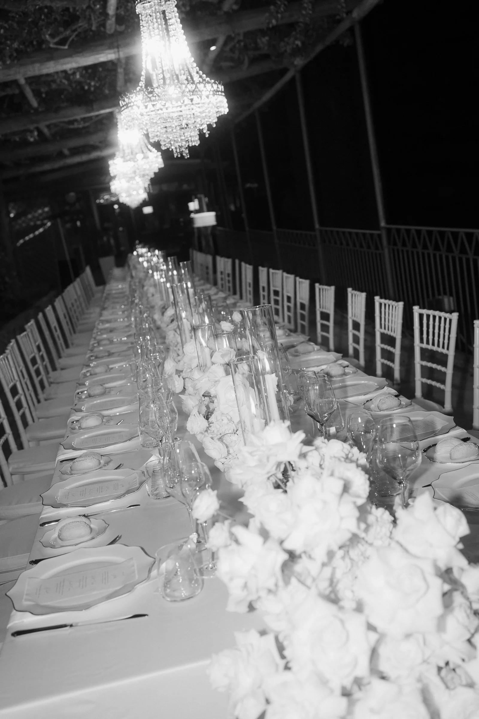 Long formal dining table with white tablecloth and elegant place settings, including wine glasses, plates, and napkins, decorated with floral centerpieces and surrounded by white chairs, set in a grand event space with chandeliers hanging from the ce