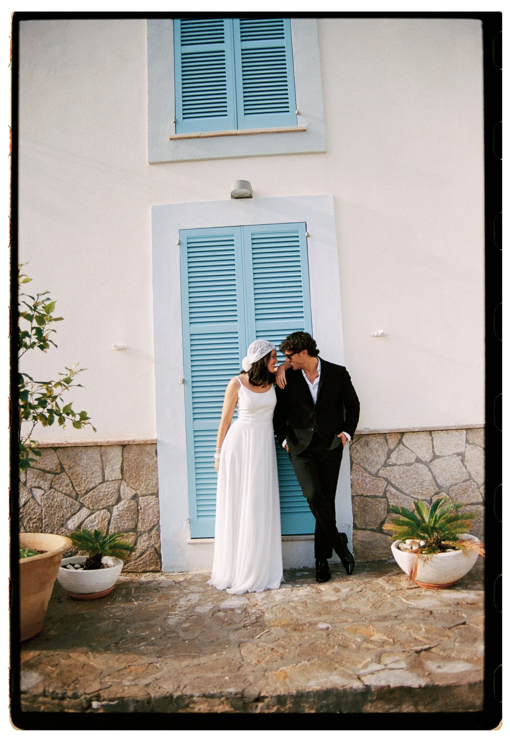 A couple in wedding attire standing in front of a white house with blue shutters, potted plants on the stone-paved ground.