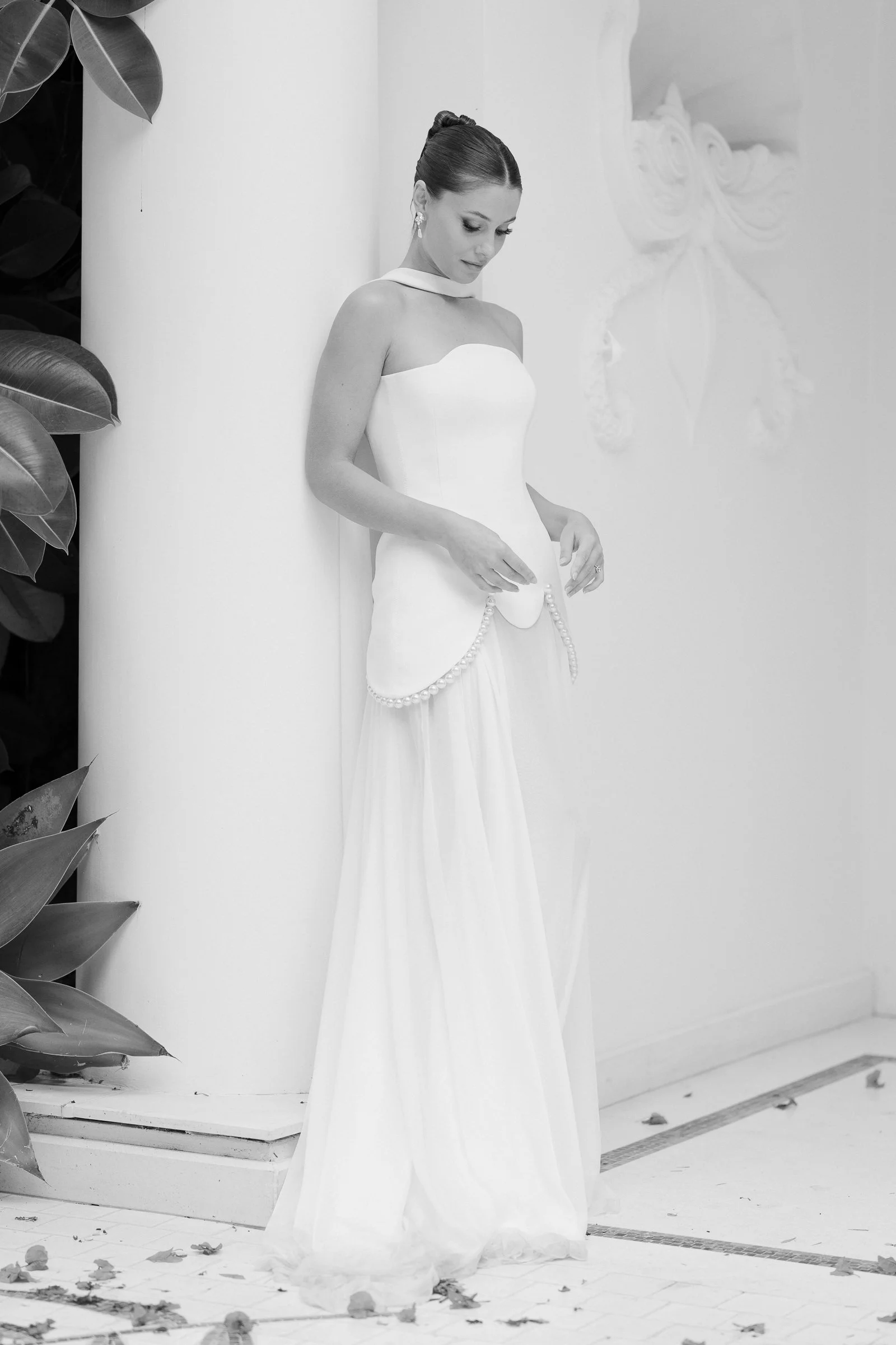 A woman in a white wedding dress standing against a white wall, with leaves and flowers scattered on the floor around her.