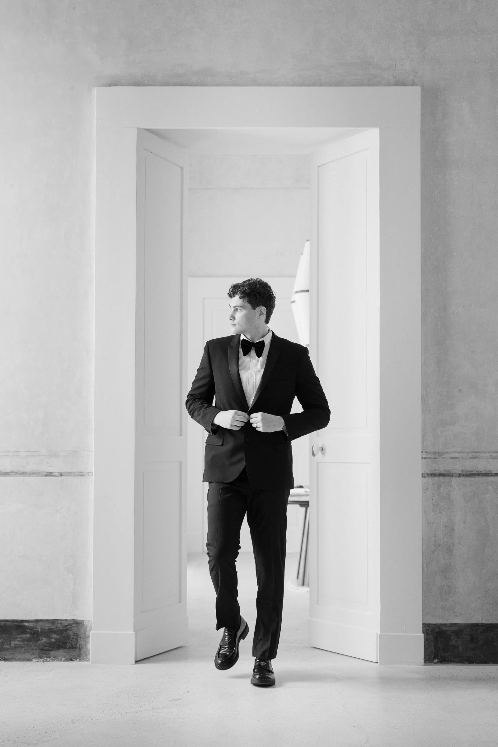 A man in a tuxedo standing in an open doorway, looking to the side with hands adjusting his jacket, in a monochrome photo.