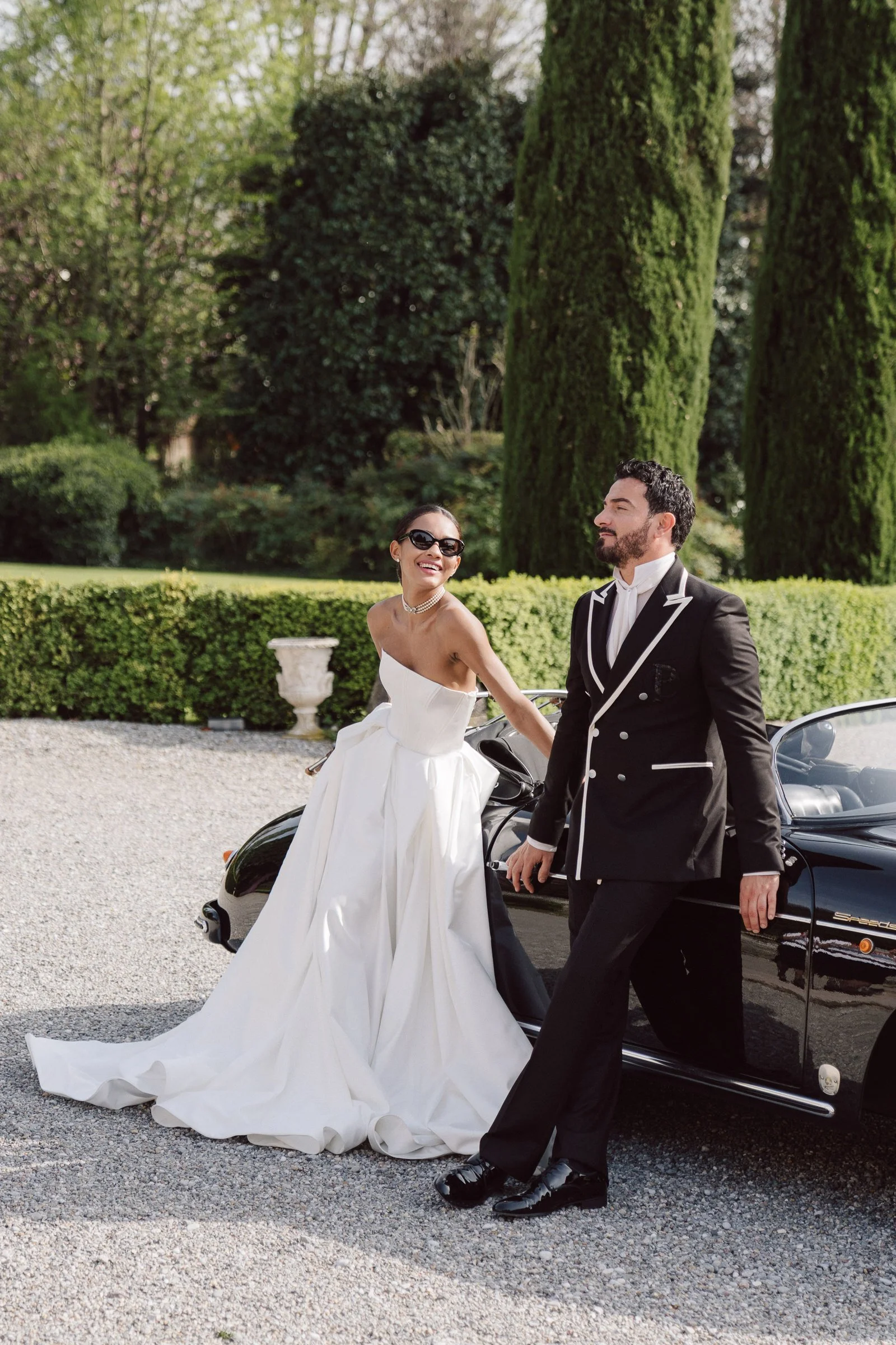 A bride and groom dressed in wedding attire standing beside a black sports car outdoors in a garden setting with green trees and bushes in the background.