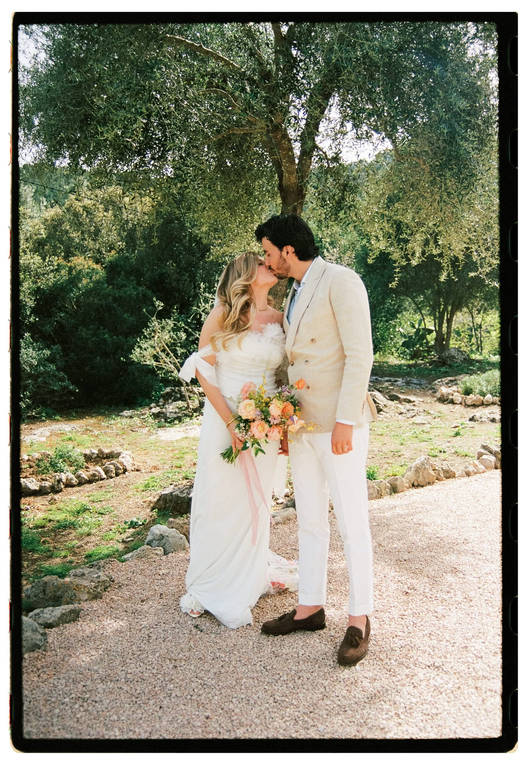 A couple dressed in wedding attire standing outdoors, kissing beneath a large tree with green foliage, with a bouquet of flowers in the bride's hand.