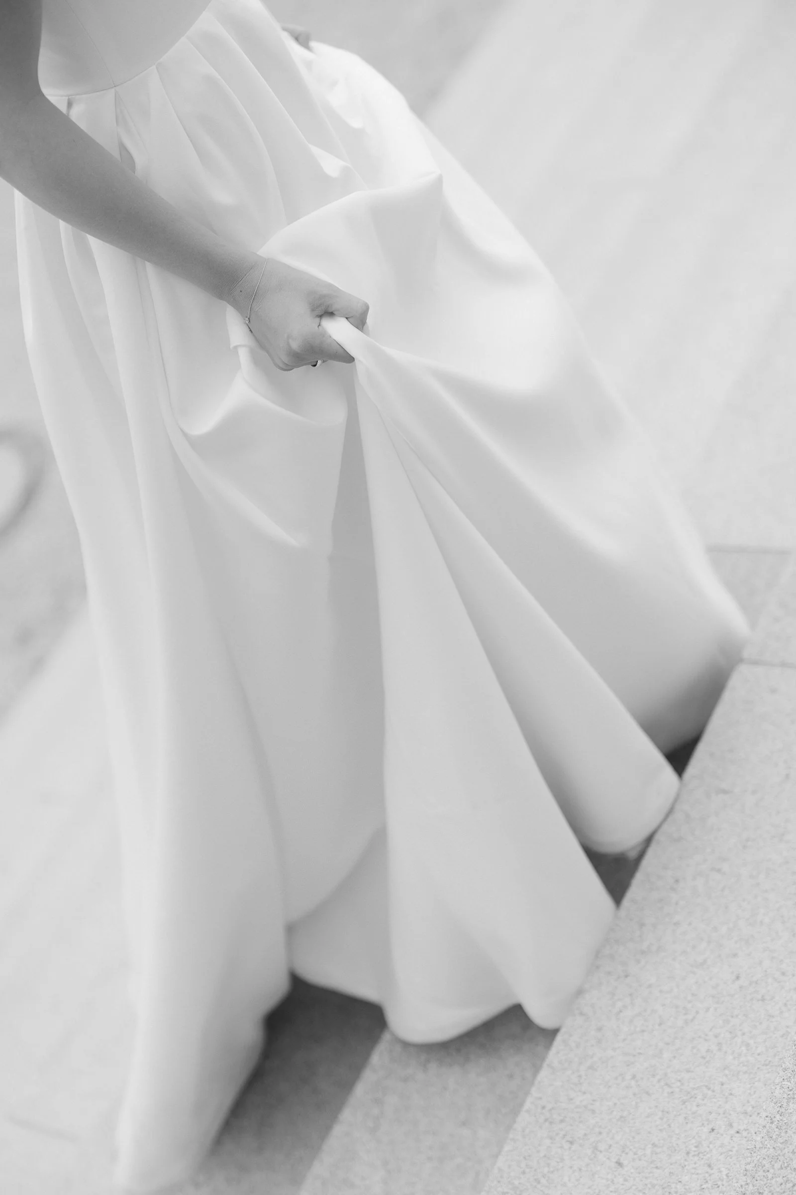 Close-up of a person holding the edge of a white gown or dress in black and white.