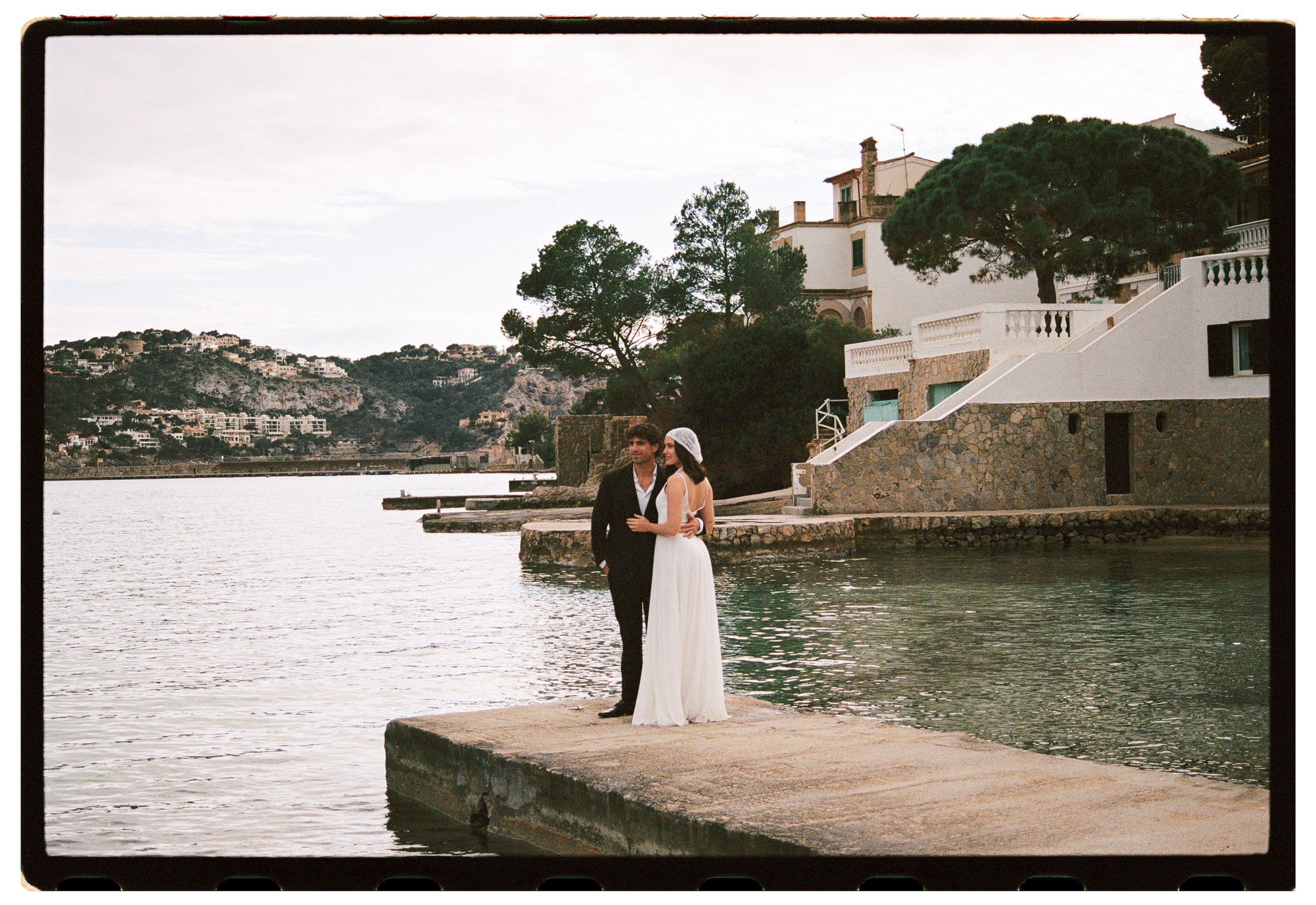 A couple dressed in formal wedding attire standing on a concrete pier by the water, with a backdrop of trees, residential buildings, and hilly landscape.