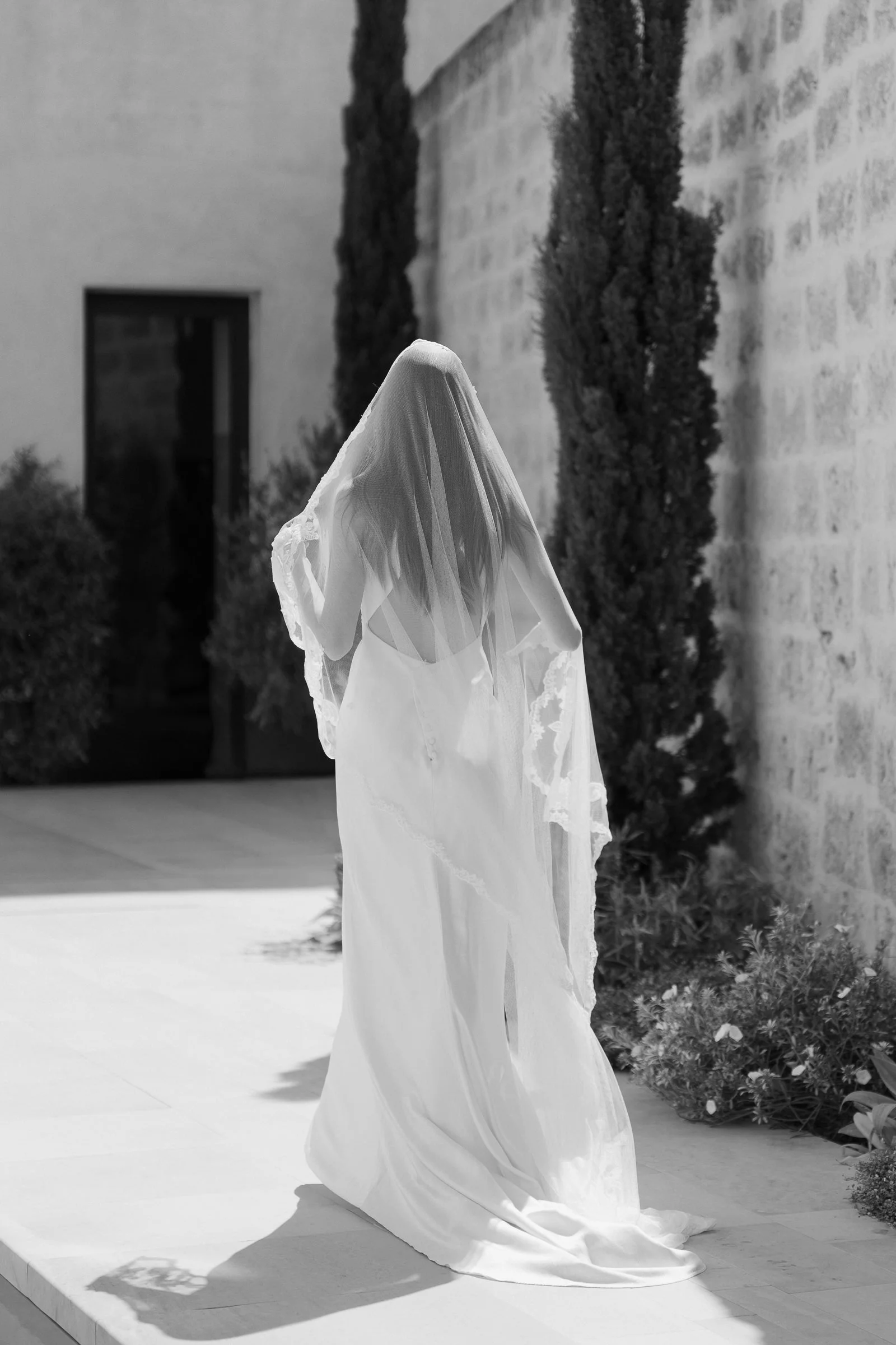 A bride wearing a flowing white wedding dress and a veil walking outside near a brick wall with tall skinny trees.