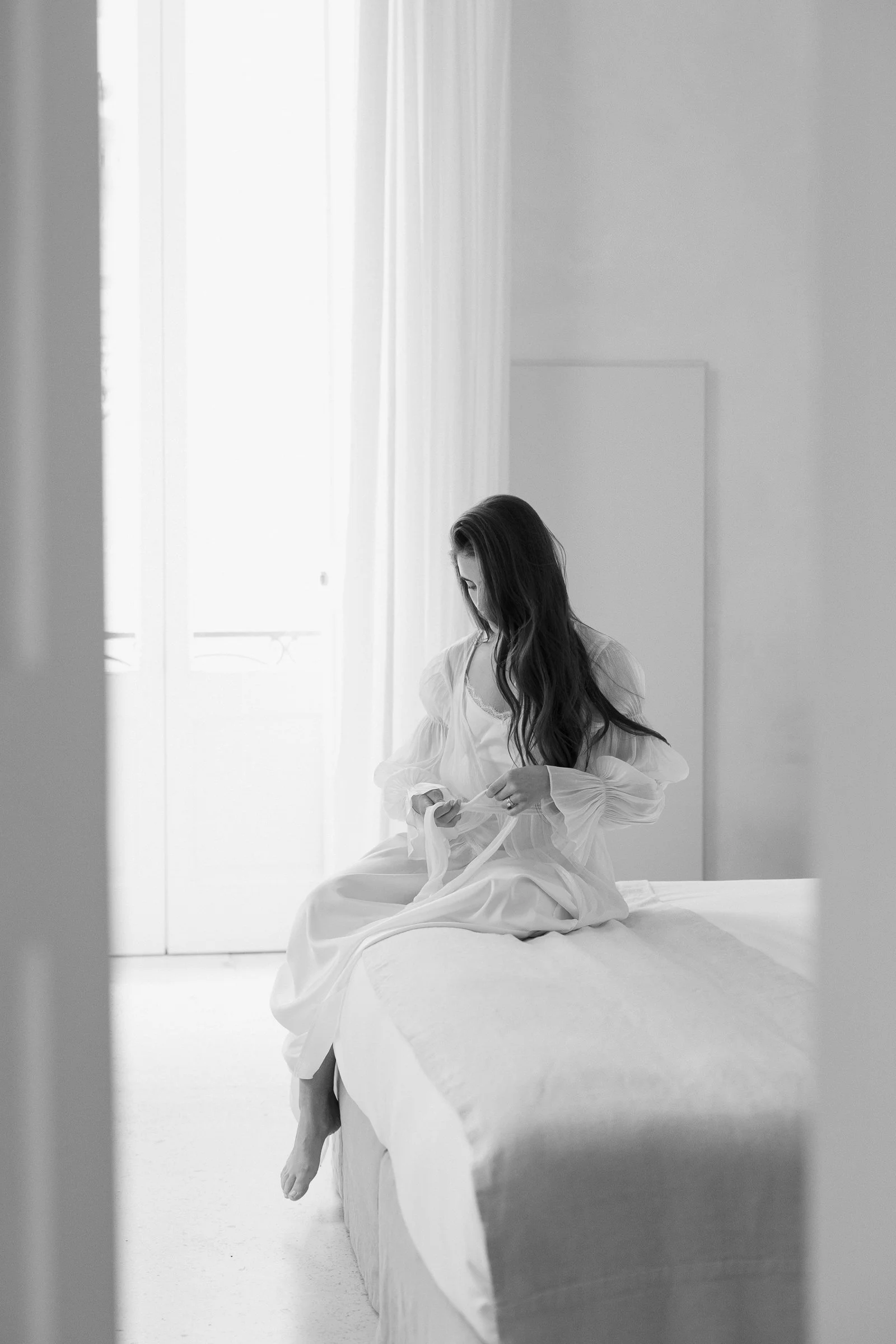 A woman sitting on a bed in a white room, looking down and holding a piece of cloth, with soft natural light coming through a window.
