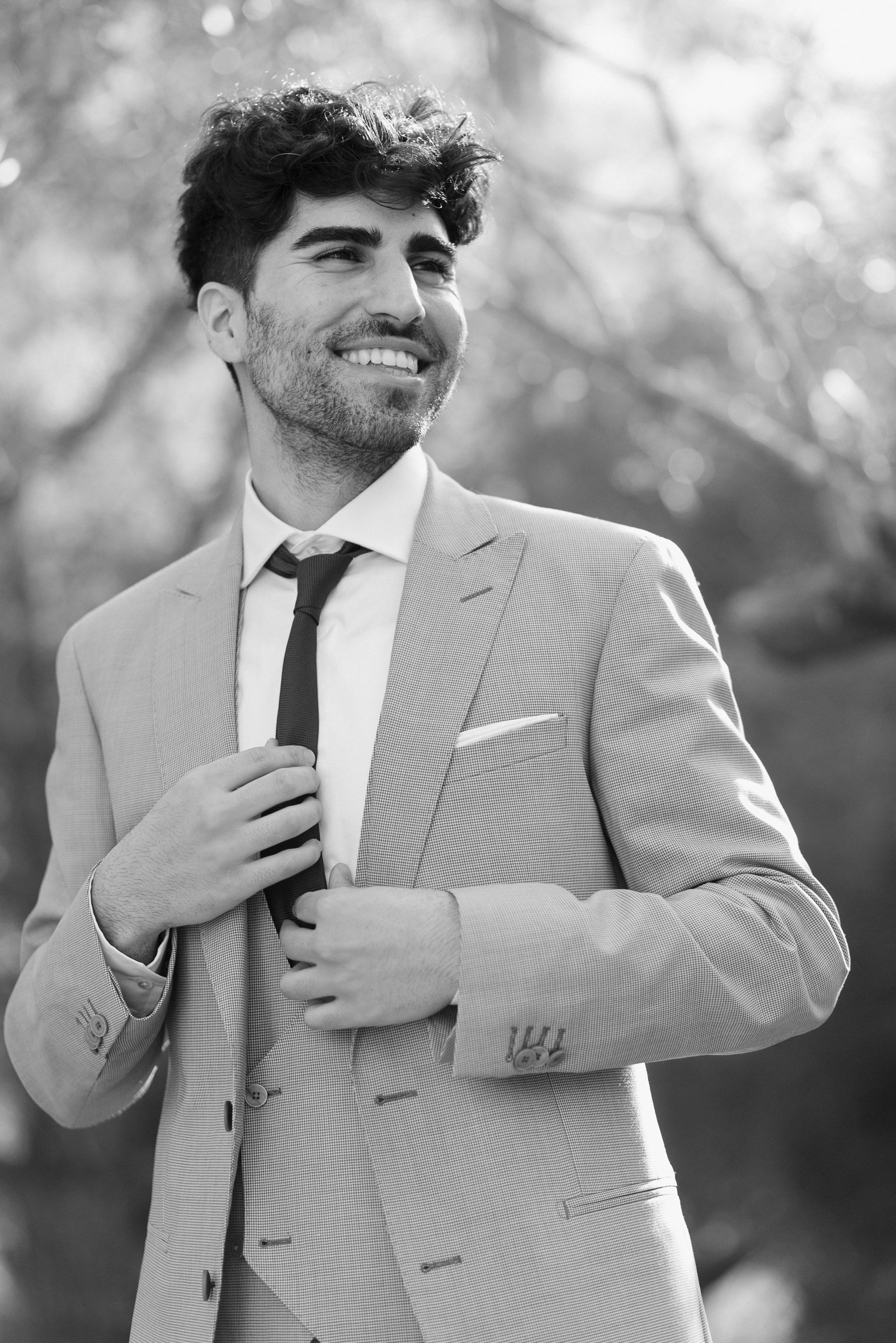 A smiling man in a light-colored suit with a dark tie, adjusting his tie, outside with blurred trees in the background.