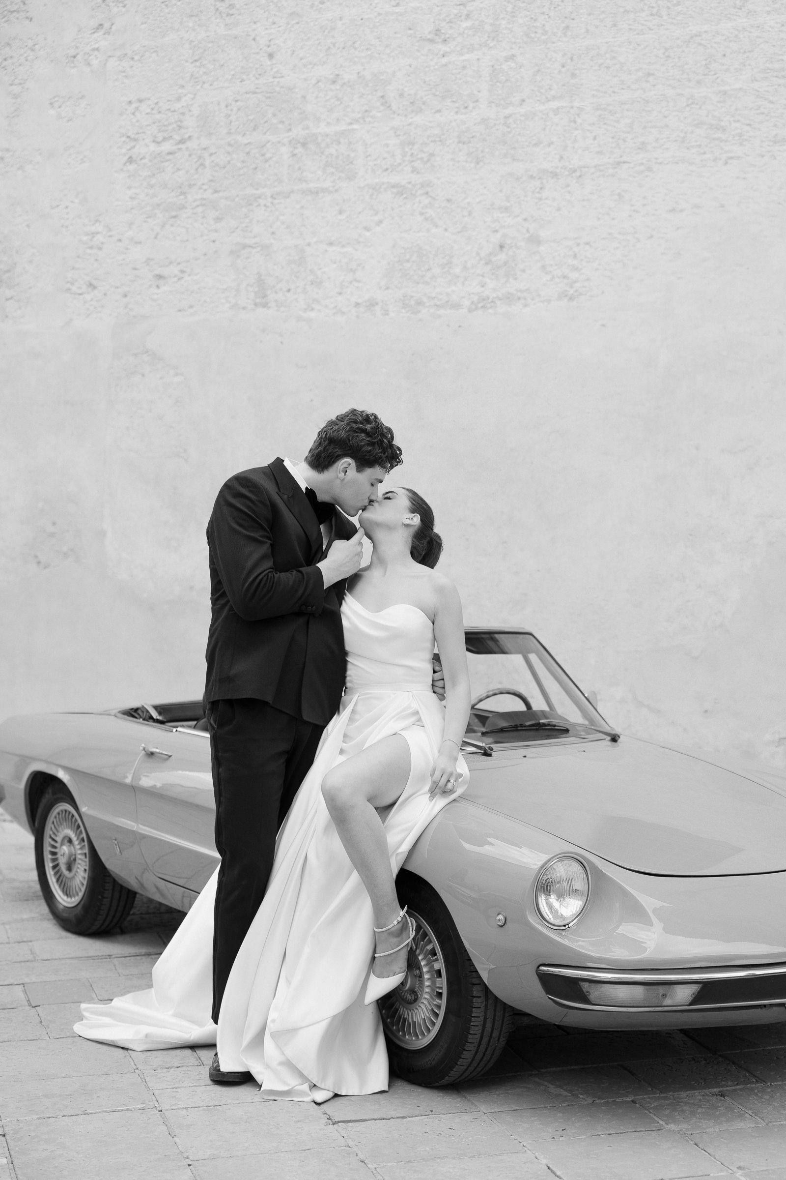 A black and white photo of a man in a tuxedo and a woman in a wedding dress kissing in front of a vintage car, with the woman resting her foot on the car's bumper.