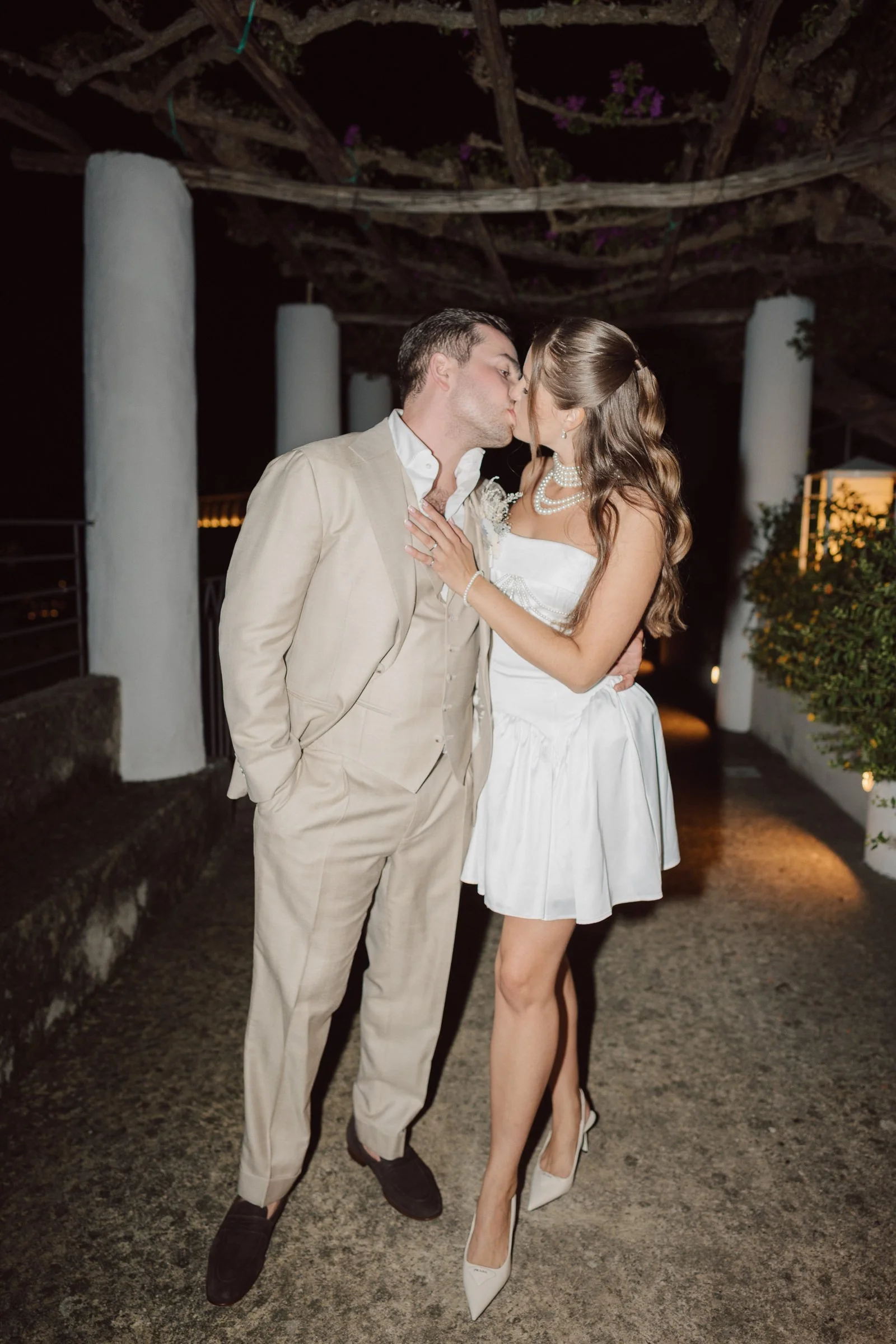 A newlywed couple sharing a kiss at night, the woman in a white dress with pearl necklaces and the man in a beige suit, standing outdoors with large white columns and a trellis with vines overhead.