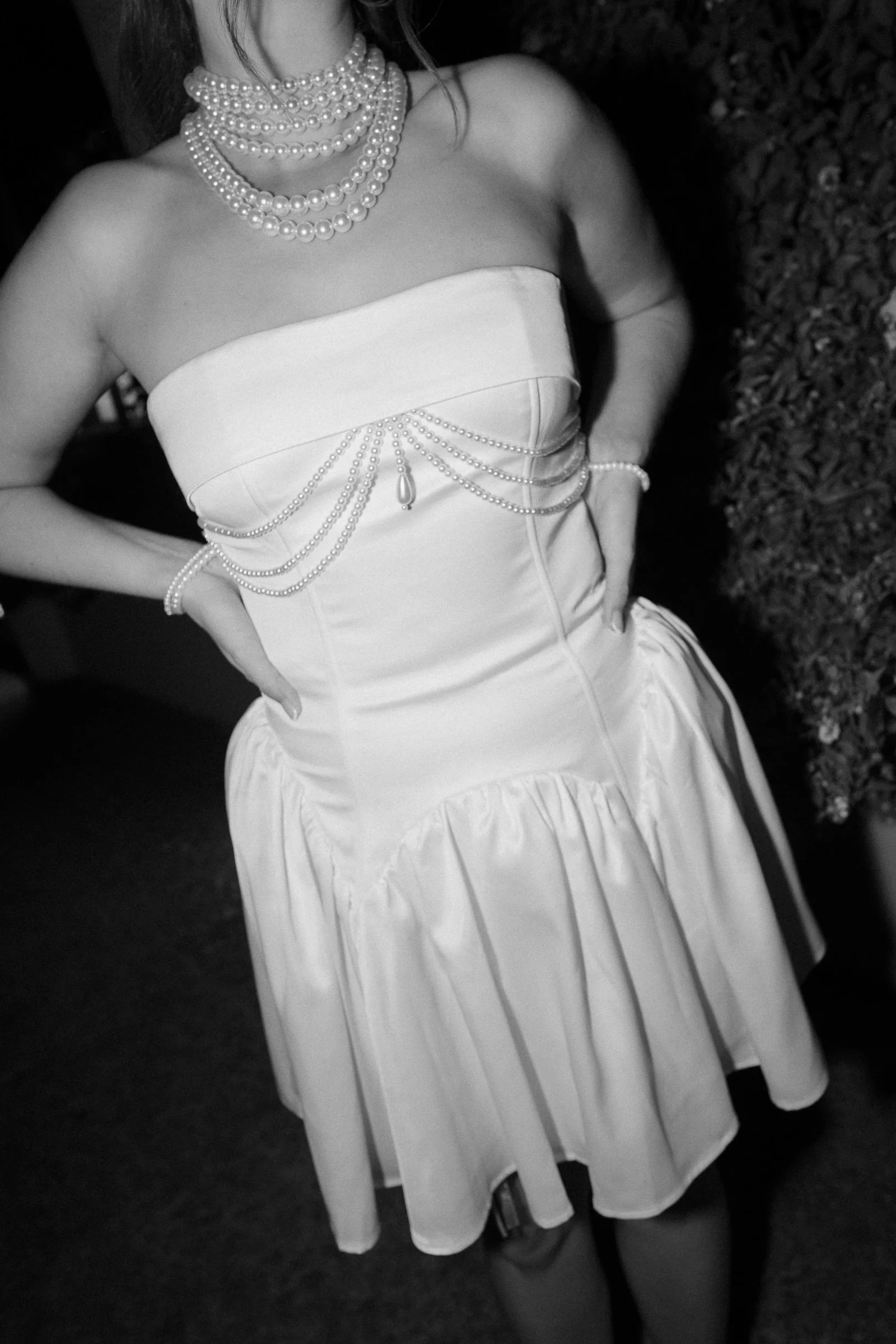 A woman wearing a strapless dress with multiple pearl necklaces and a pearl bracelet, standing outdoors on grass.