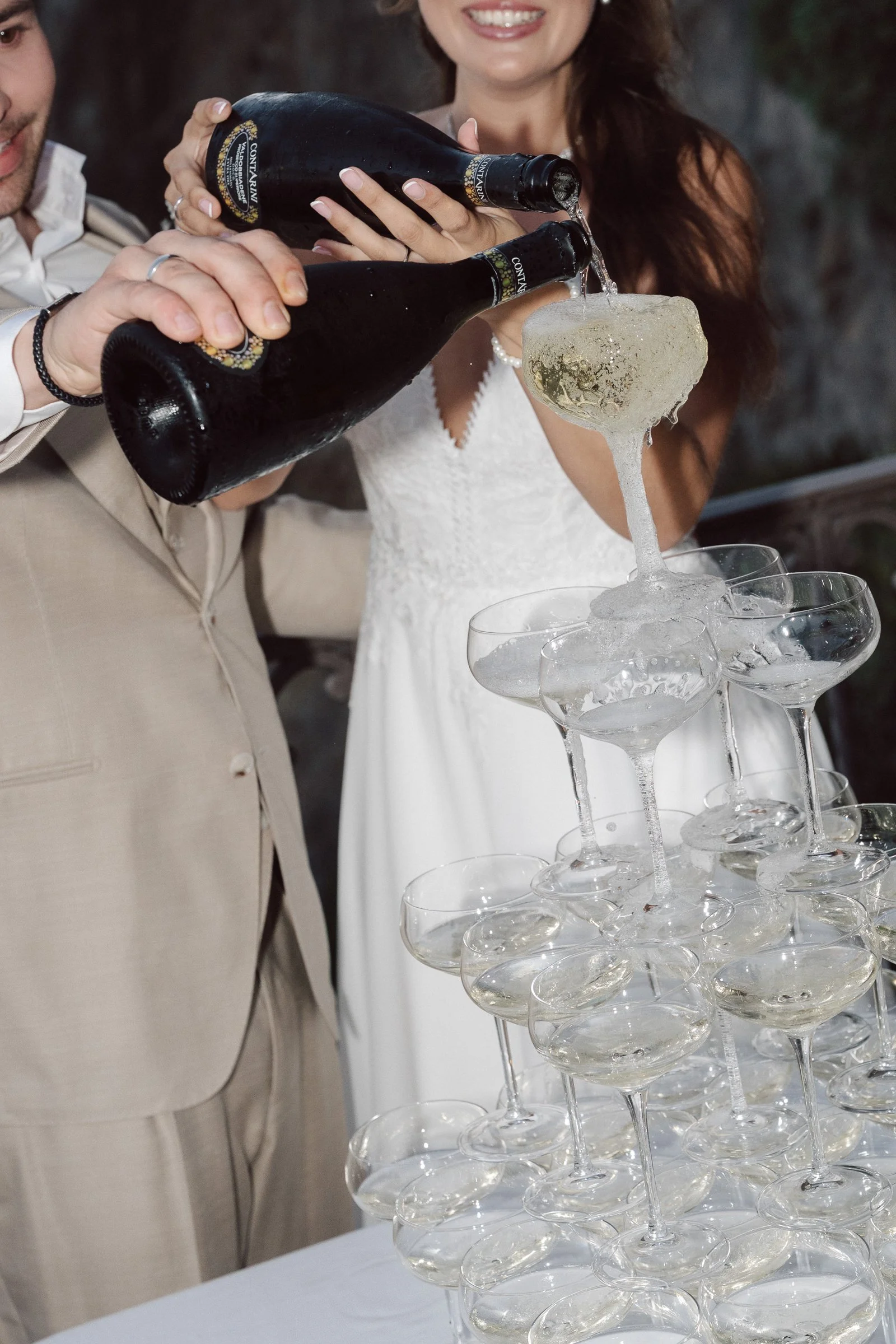 A couple dressed in formal attire pouring champagne into a tower of glasses at a celebration event.