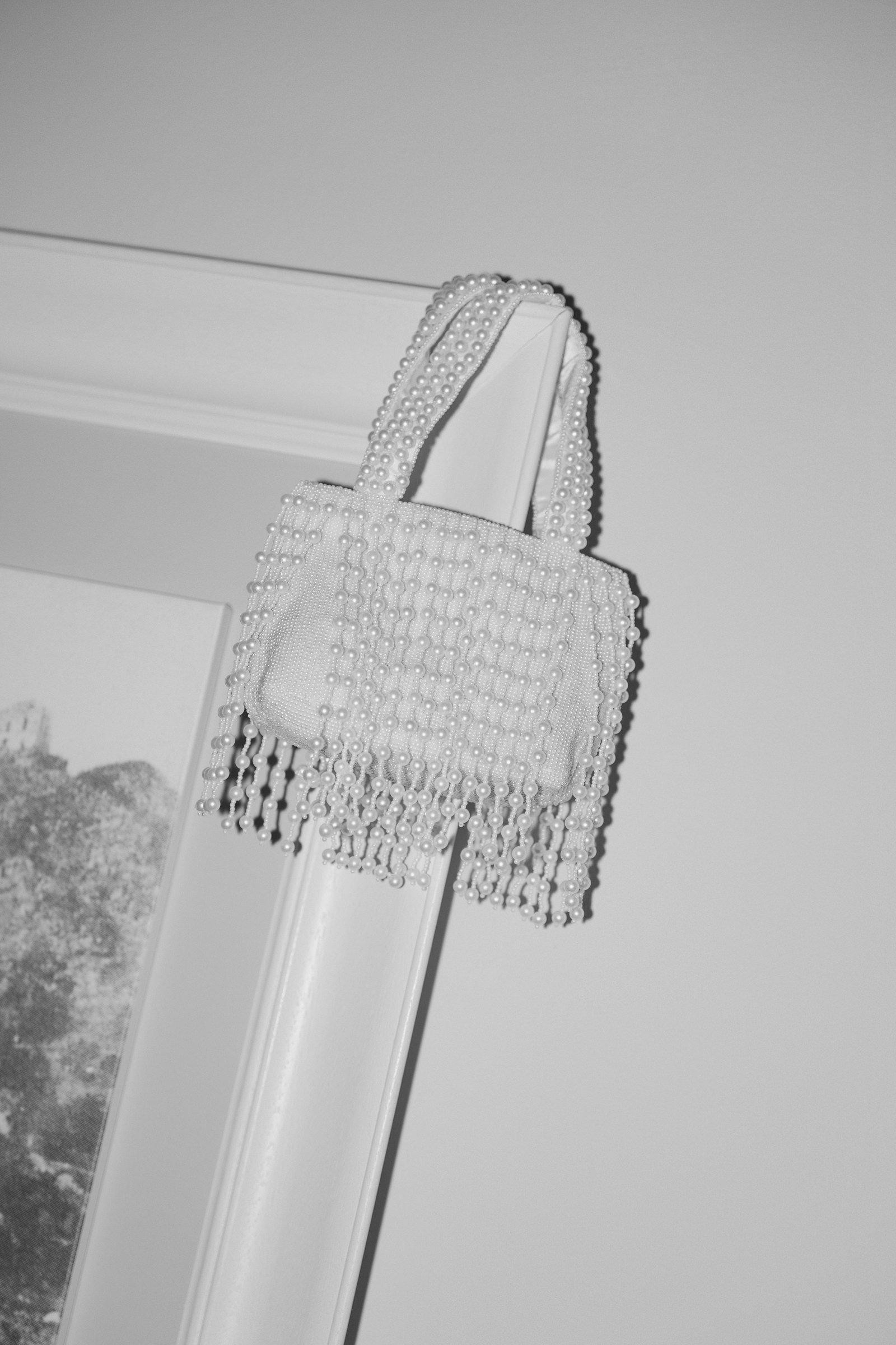 A pearl-embellished handbag hanging from a door trim with a pearl strap, black and white photo.