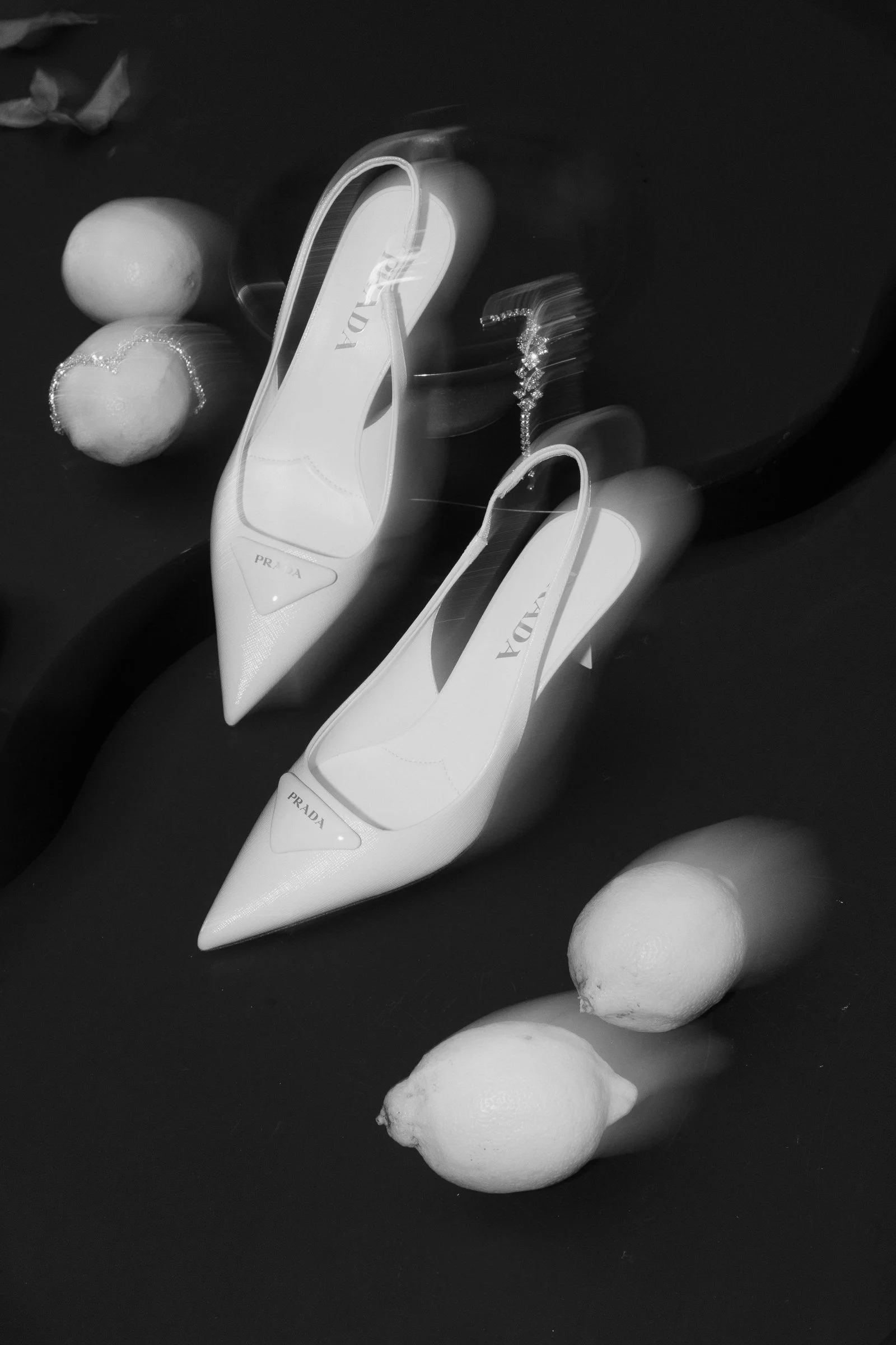 Black and white photo of two white Prada high-heeled shoes placed on a dark surface, with several round objects arranged around them, appearing to be meringue or similar confections.
