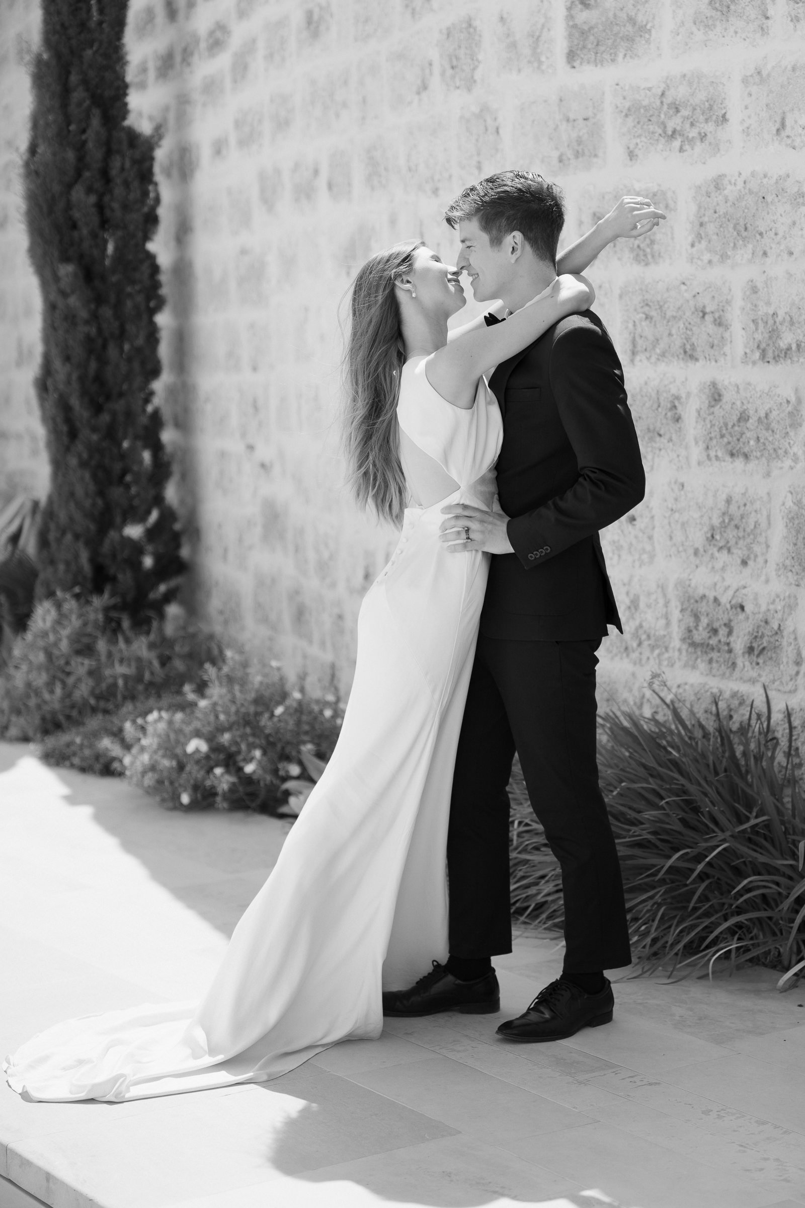 A black-and-white photograph of a couple in wedding attire, celebrating outdoors. The woman is wearing a white wedding dress and the man is in a black suit and bow tie. They are standing close, smiling, with their noses touching, in front of a textur